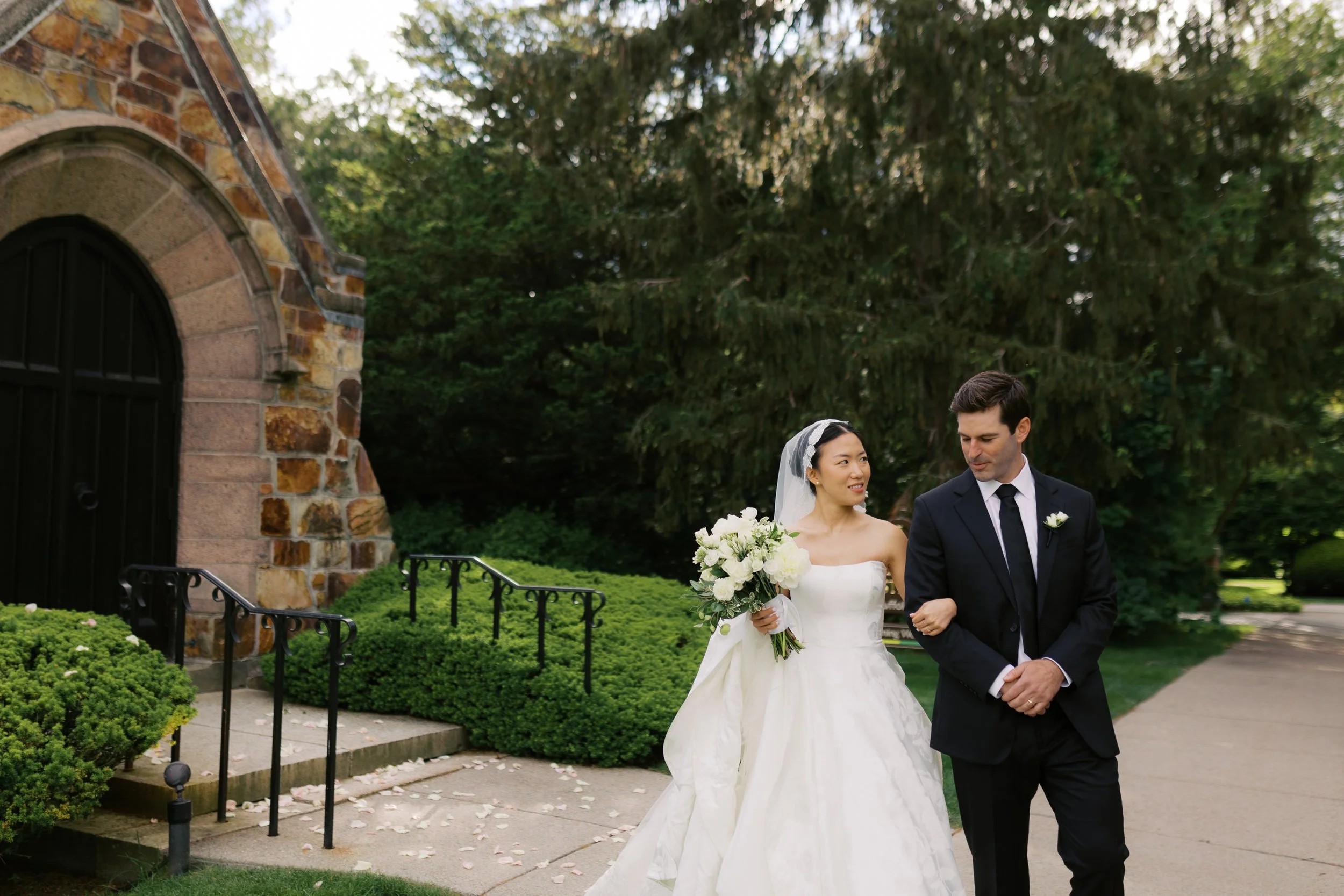 A bride in a white wedding gown and veil holding a bouquet, walking arm-in-arm with a groom in a dark suit, outside in a park-like setting.