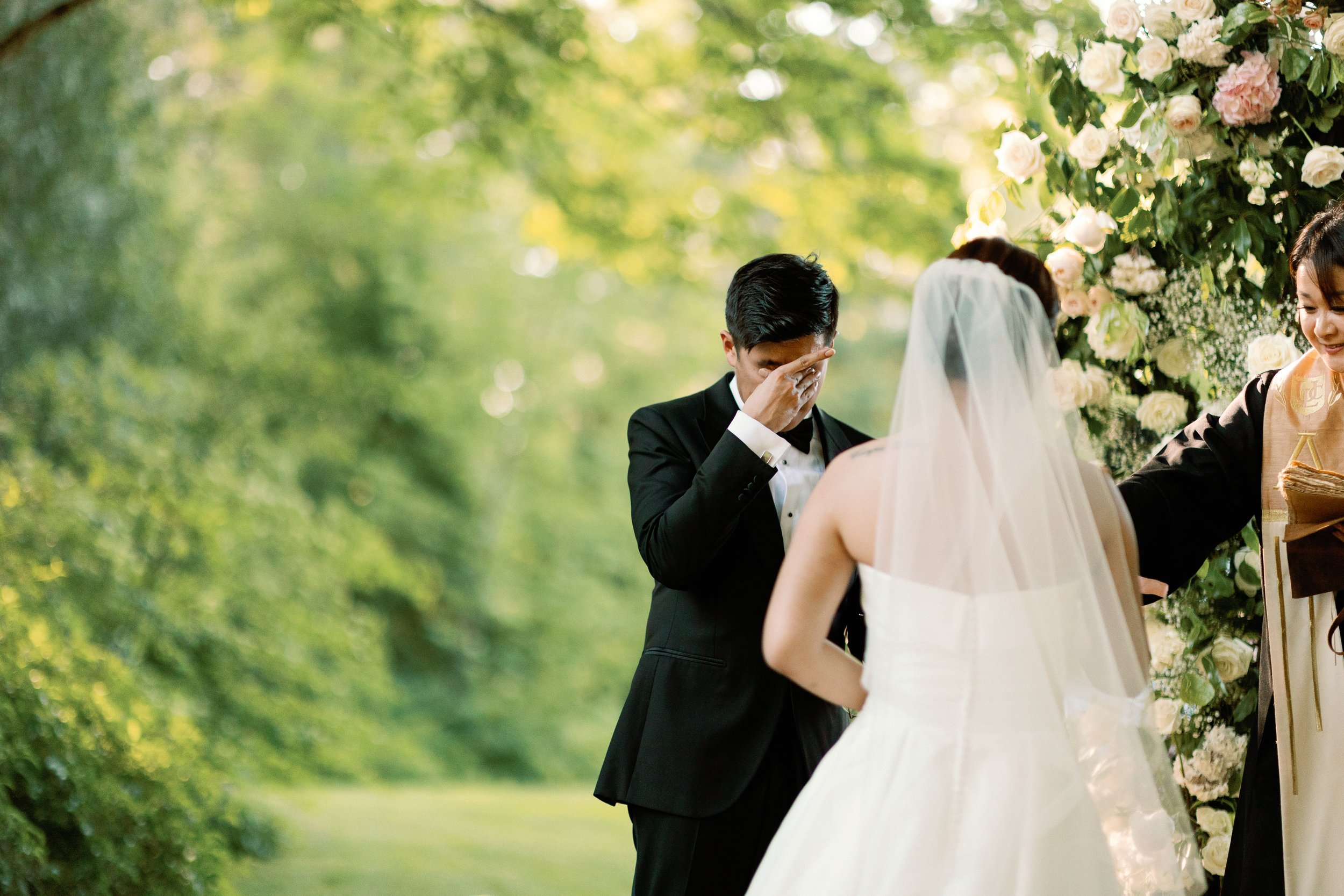 A groom is emotional during his outdoor wedding ceremony, covering his face with his hand, with a bride facing him and a floral arch in the background.