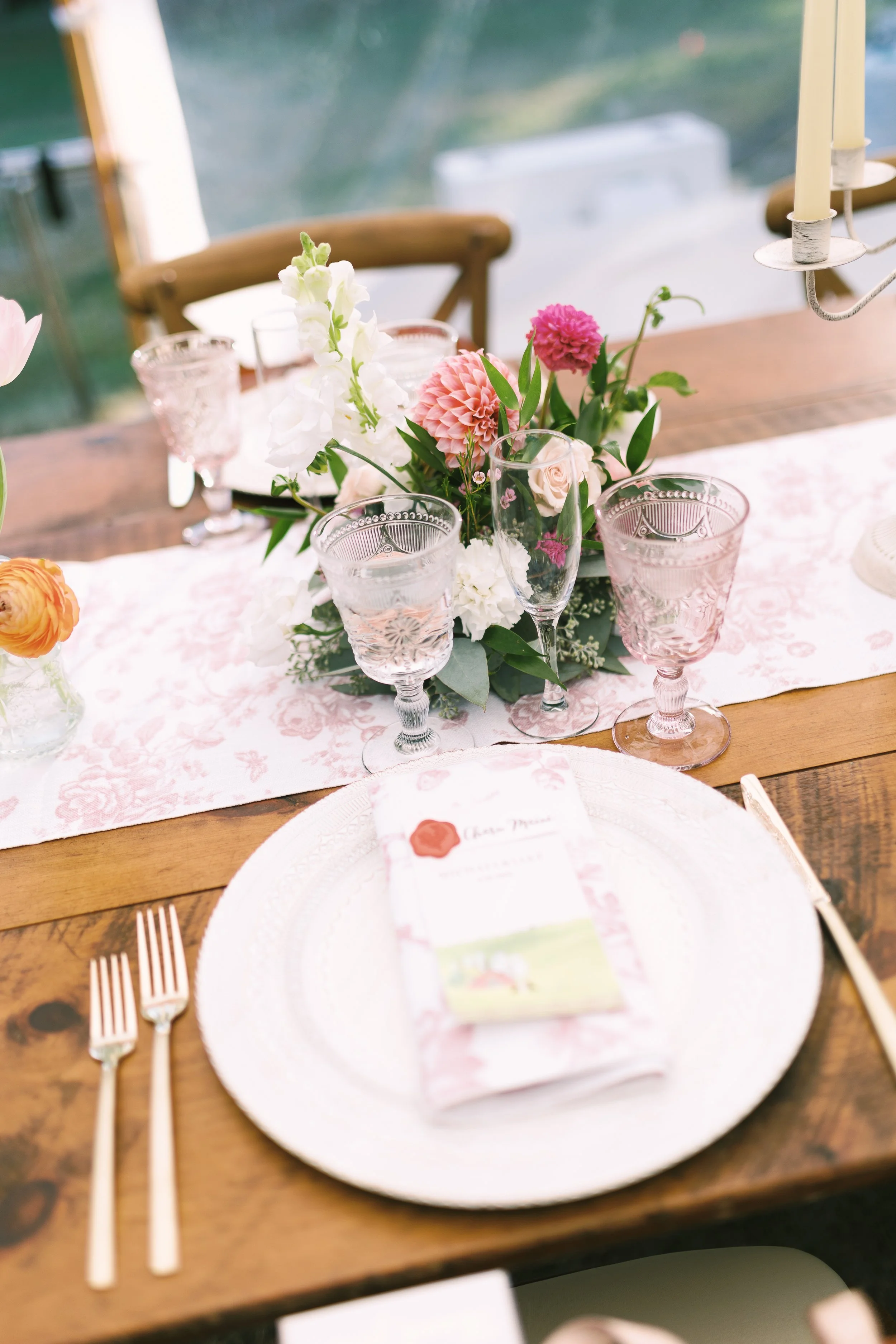 A decorated table with a floral centerpiece, pink and clear glassware, a white plate with a folded paper napkin, gold forks, and a white and pink table runner with a floral pattern.