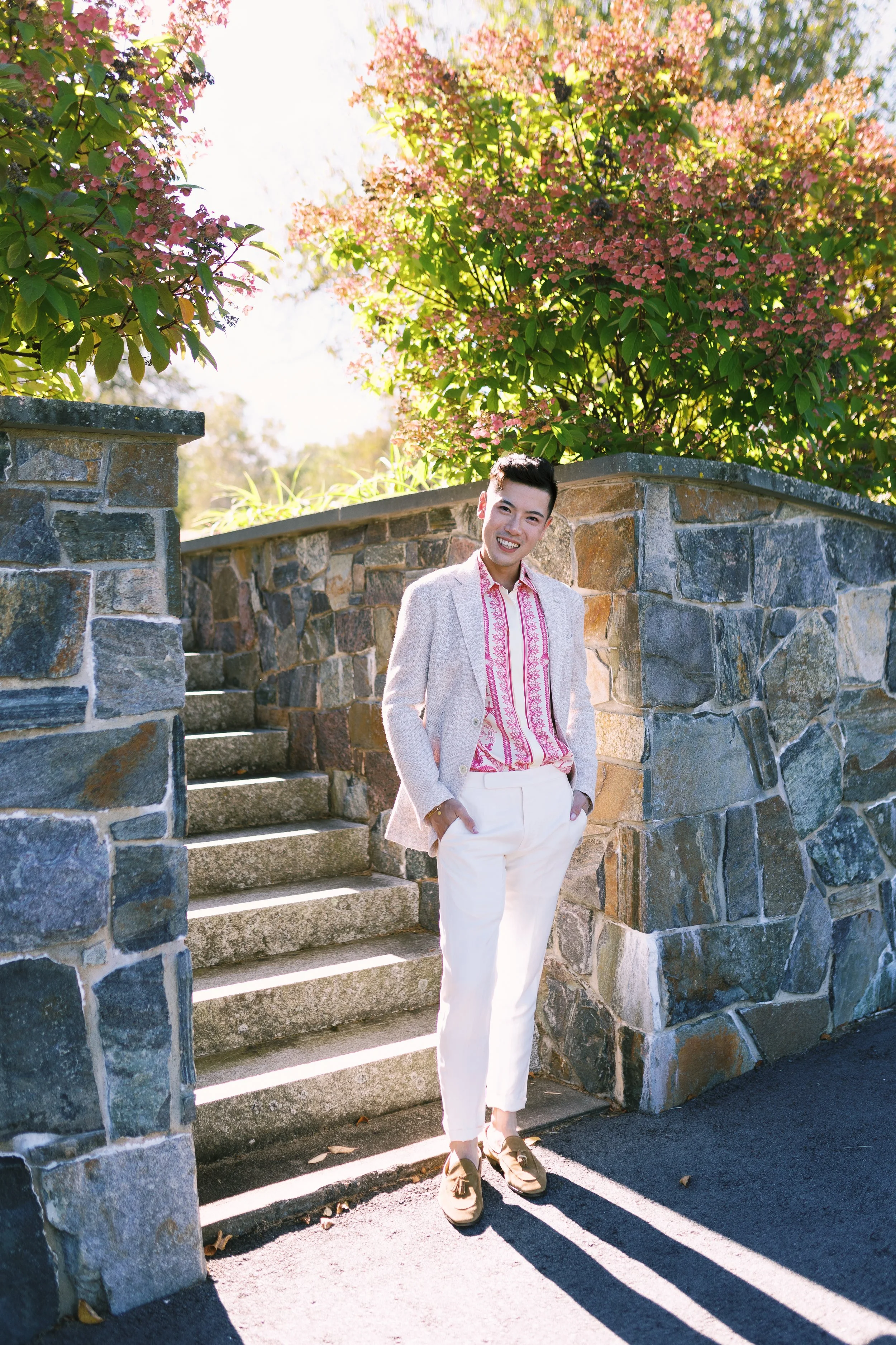 A young man in a beige blazer, patterned pink and white shirt, white pants, and brown shoes standing outdoors on a sunny day. He is smiling with hands in pockets, near stone steps and a stone wall with lush green trees and pink flowering bushes in th