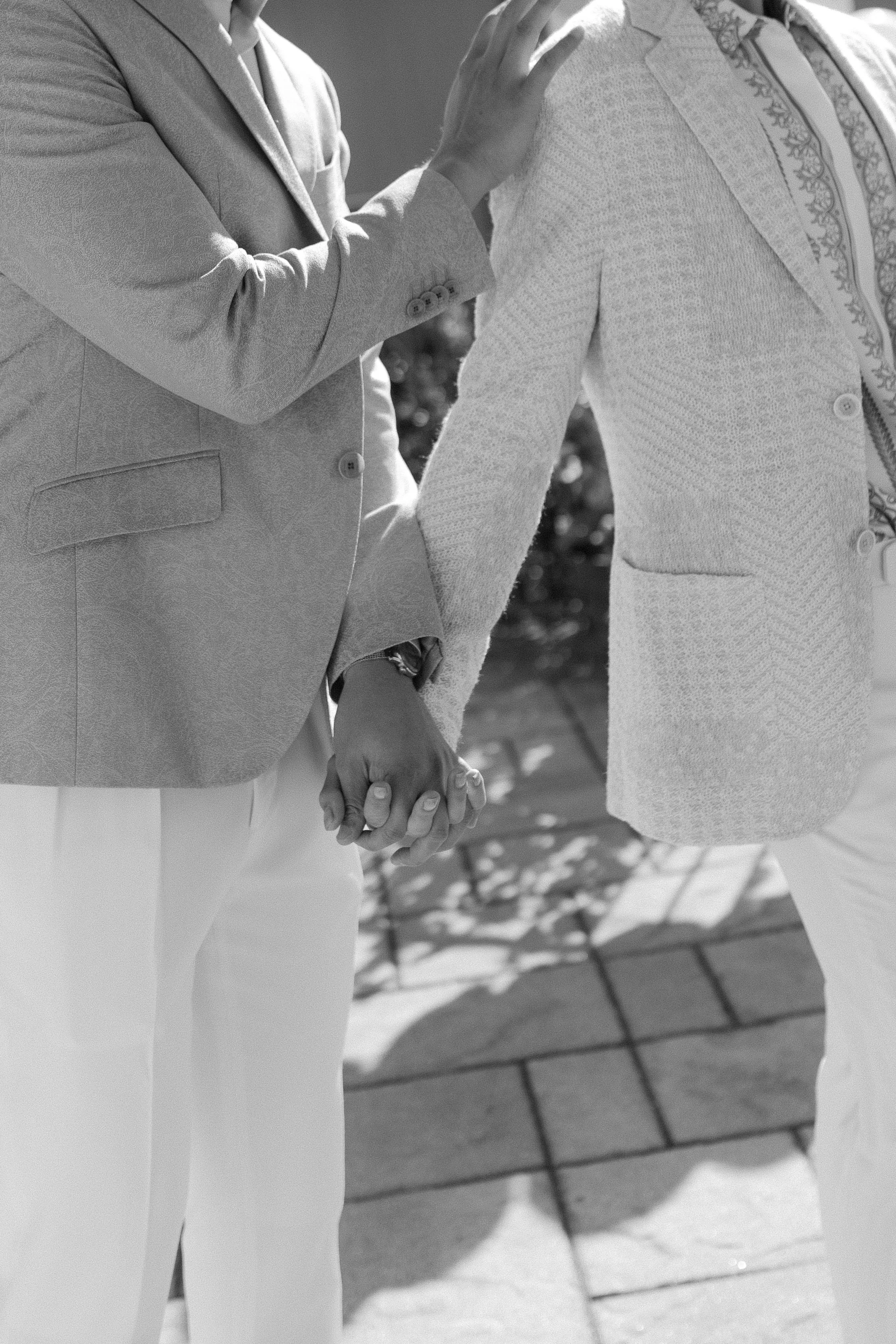 Two people holding hands, dressed in light-colored suits, outdoors on a sunny day.