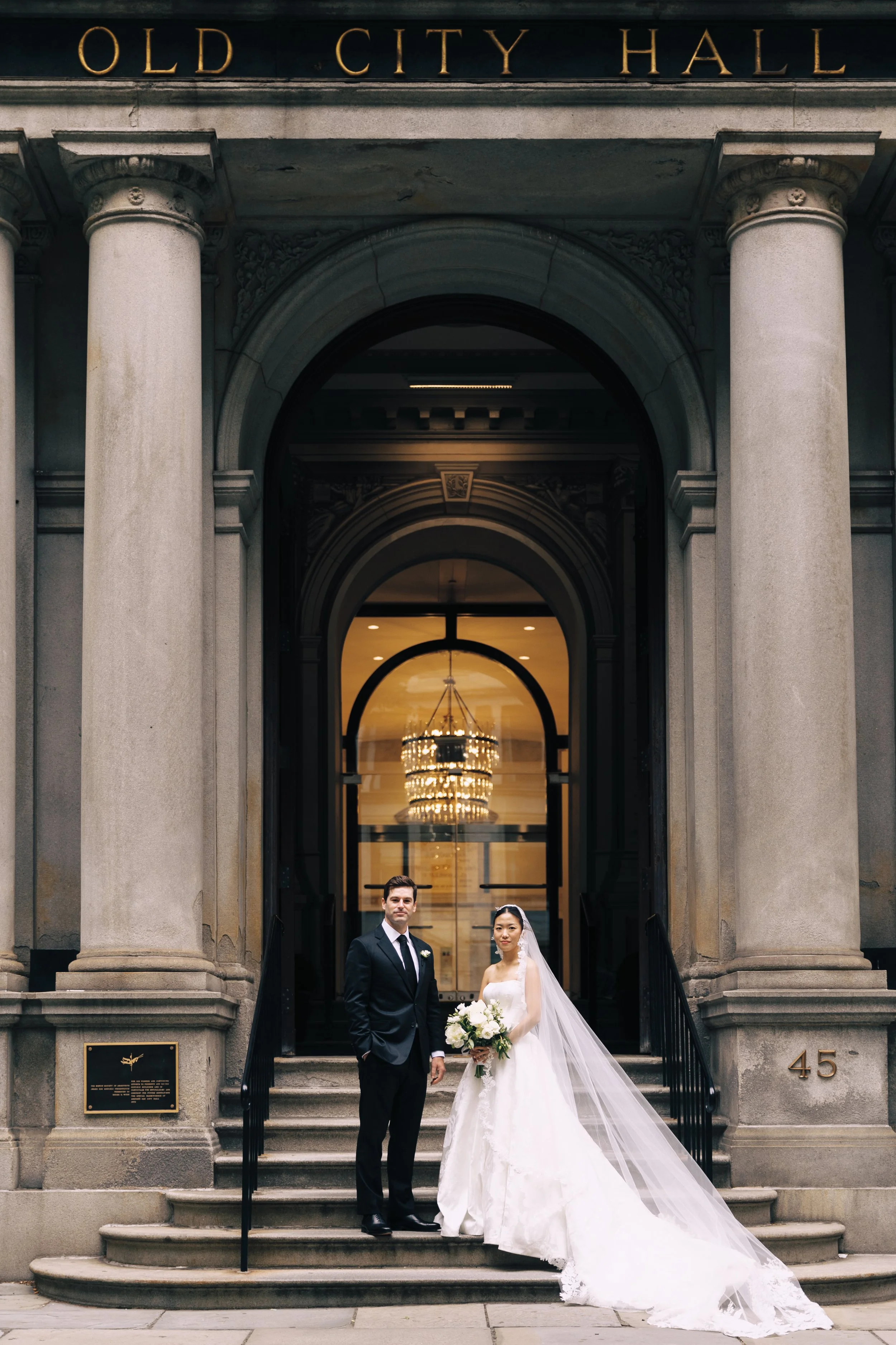 Bride and groom standing on stairs outside Old City Hall, with bride dressed in a white wedding gown holding a bouquet, and groom in a black suit and tie. The building has large columns and a chandelier visible through the glass door.