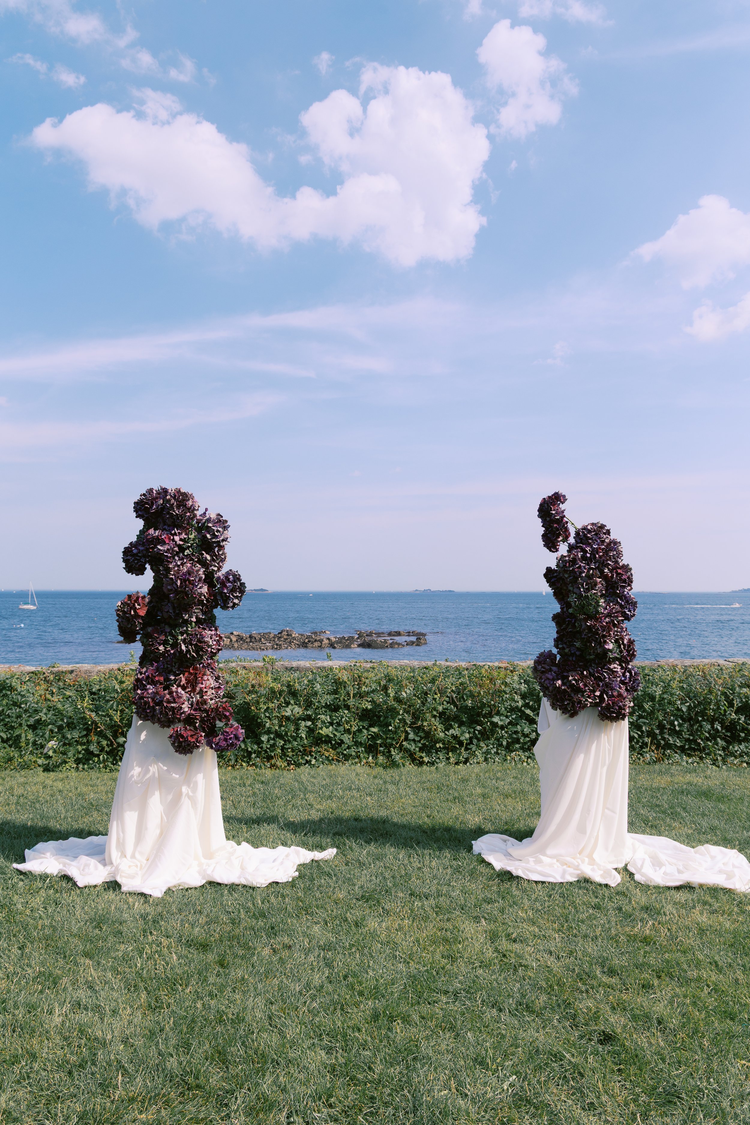 Two floral arrangements with dark purple flowers and white fabric bases on a grassy area by the ocean, with a blue sky and clouds overhead.