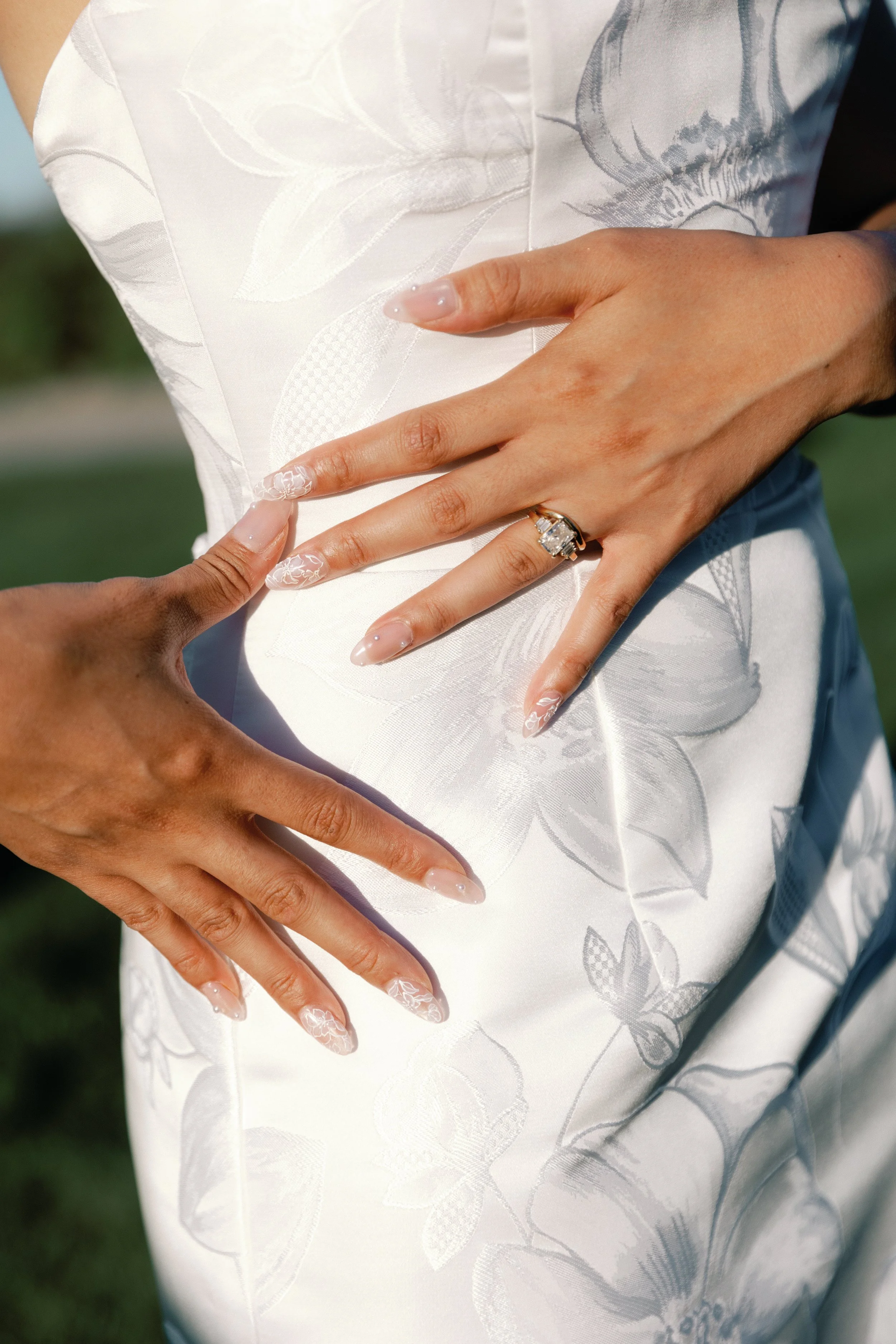 Close-up of a person wearing a white floral-patterned dress, showing two hands resting on the waist and a diamond ring on the ring finger of the right hand. The background is blurred outdoors.