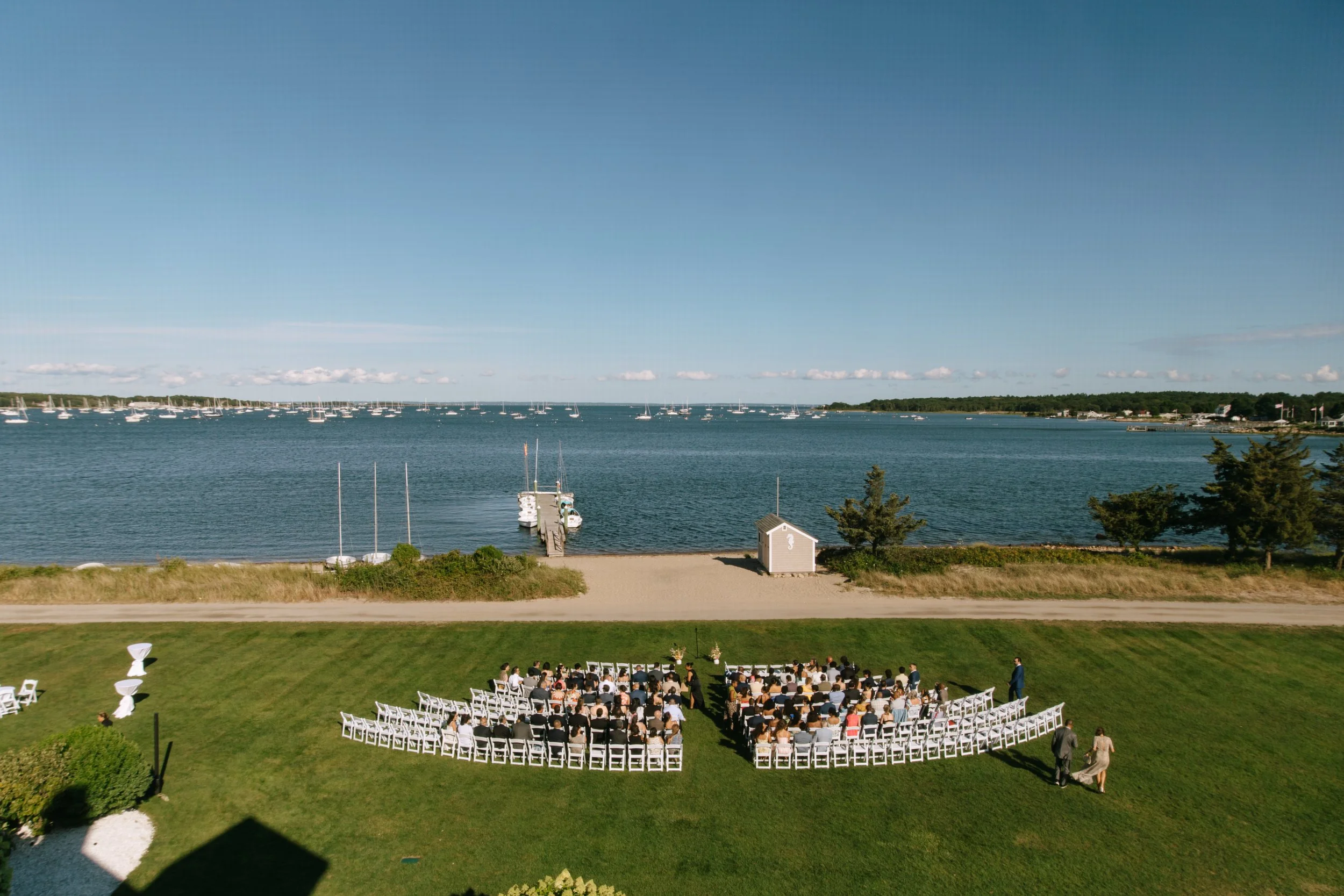 Outdoor wedding ceremony with guests seated on white chairs on green grass, overlooking a lake with sailboats and a small dock, under a clear blue sky.