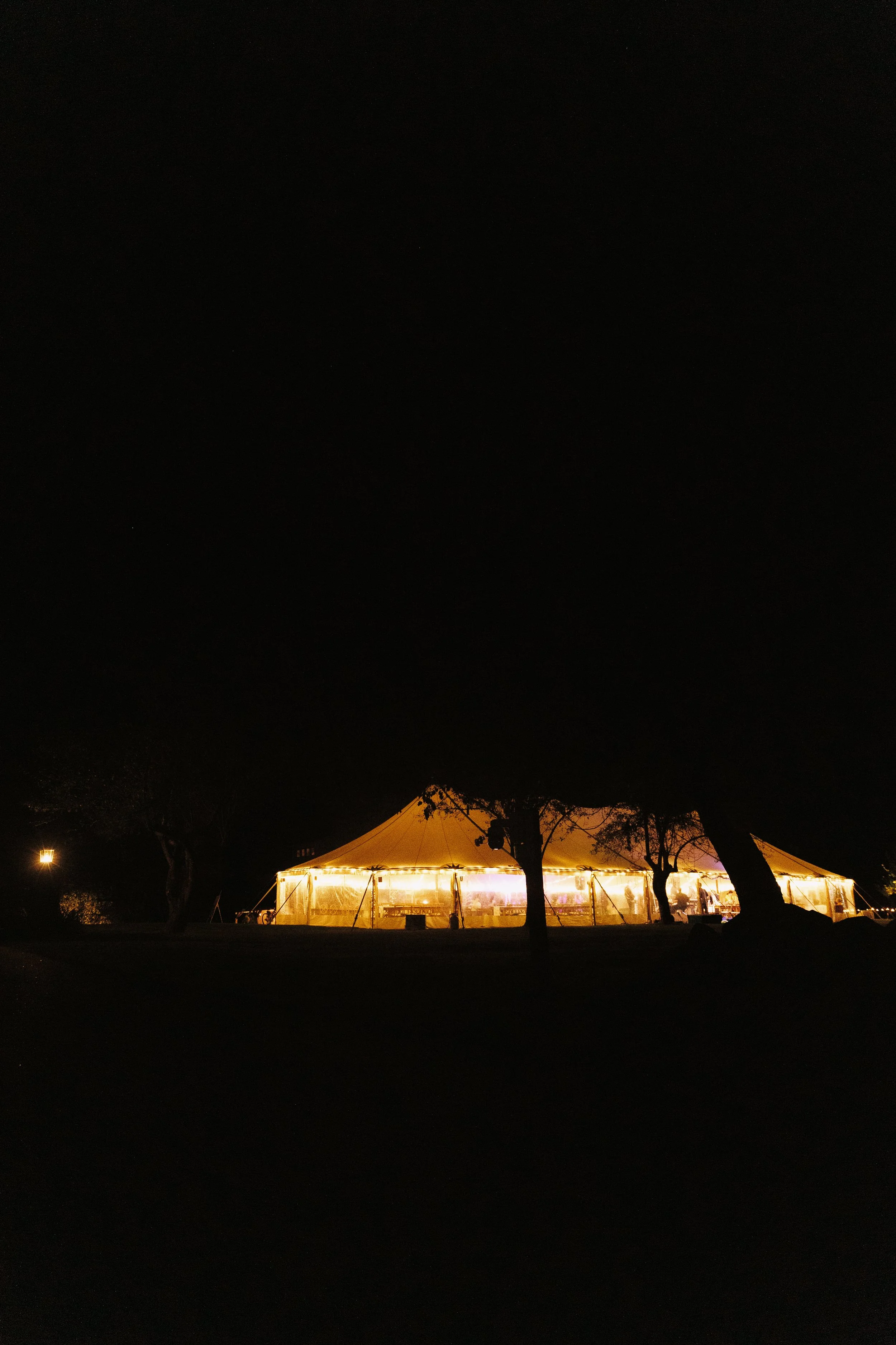 Large outdoor party tent illuminated with string lights at night, surrounded by trees and open space.