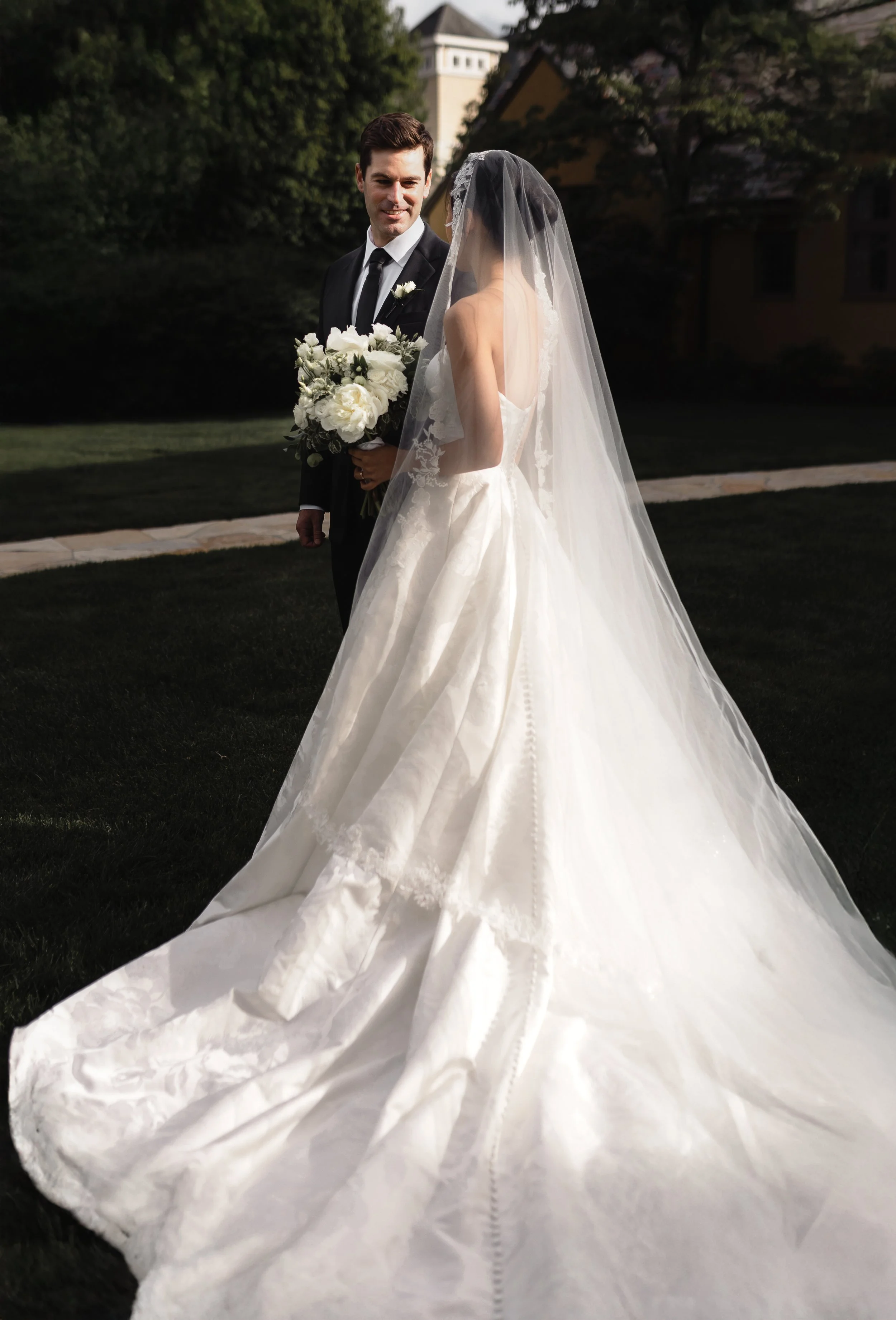 A bride and groom standing outdoors on a lawn, with the bride holding a bouquet of white flowers. The bride is wearing a white wedding gown with a long train and a veil, while the groom is in a dark suit with a white shirt and black tie.