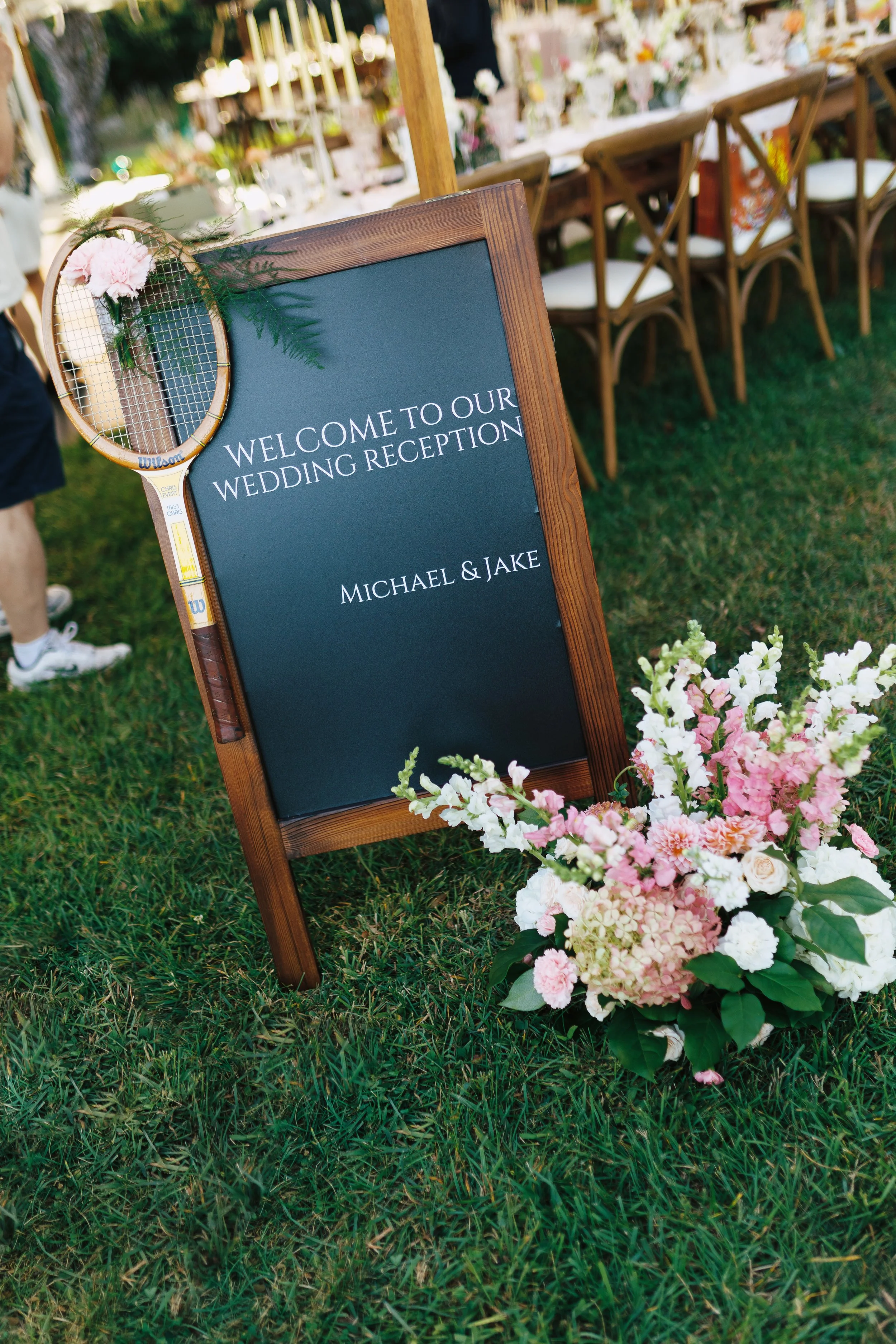 A chalkboard sign with a wooden frame greeting guests at a wedding reception, decorated with pink and white flowers, and a tennis racket with a pink flower attached hangs on the top left corner. The sign reads, "Welcome to our wedding reception, Mich