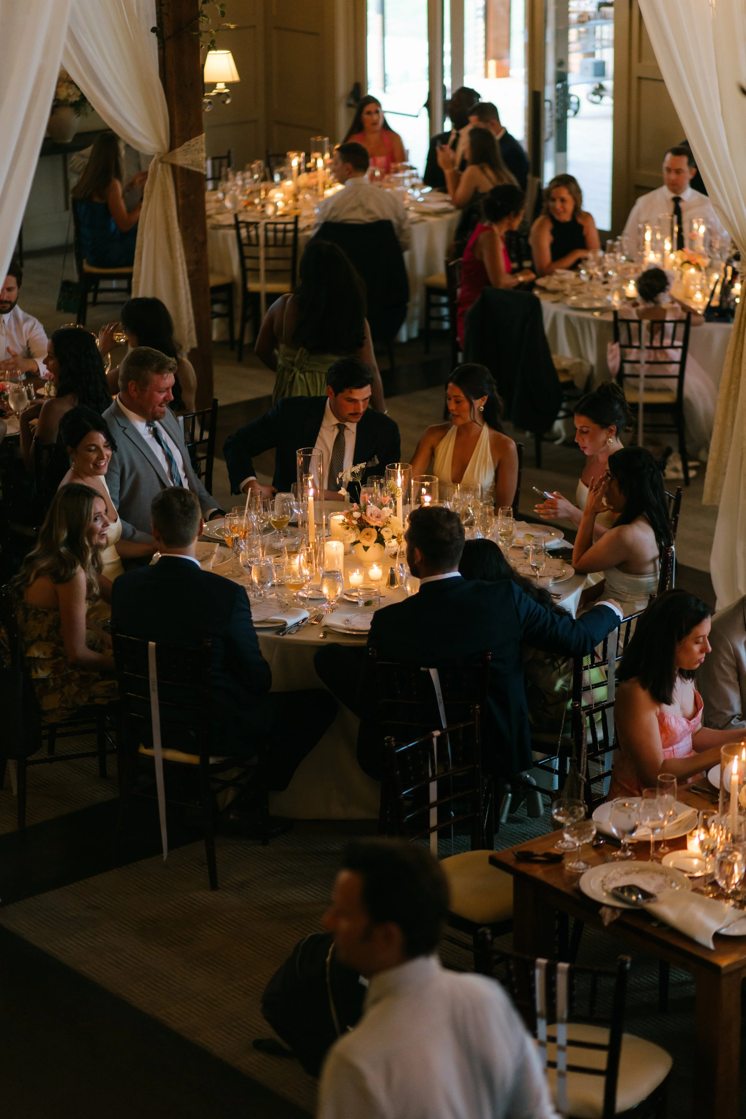 A formal indoor wedding reception with guests seated at round tables, candles, and floral centerpieces, during evening time.