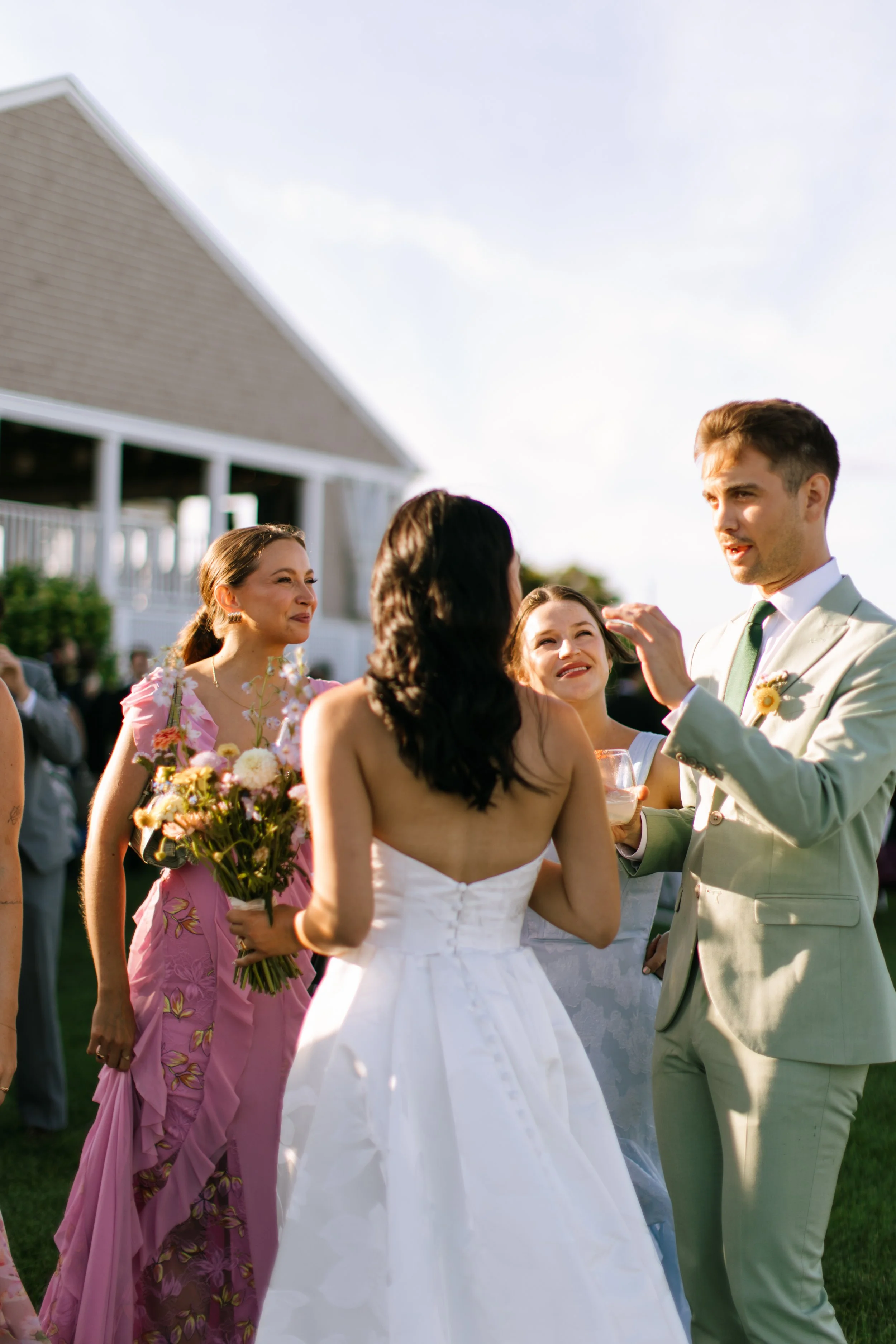 A wedding reception outdoors with people celebrating, including a bride in a white dress, a groom in a light-colored suit, and women holding flowers, with a house and a clear sky in the background.