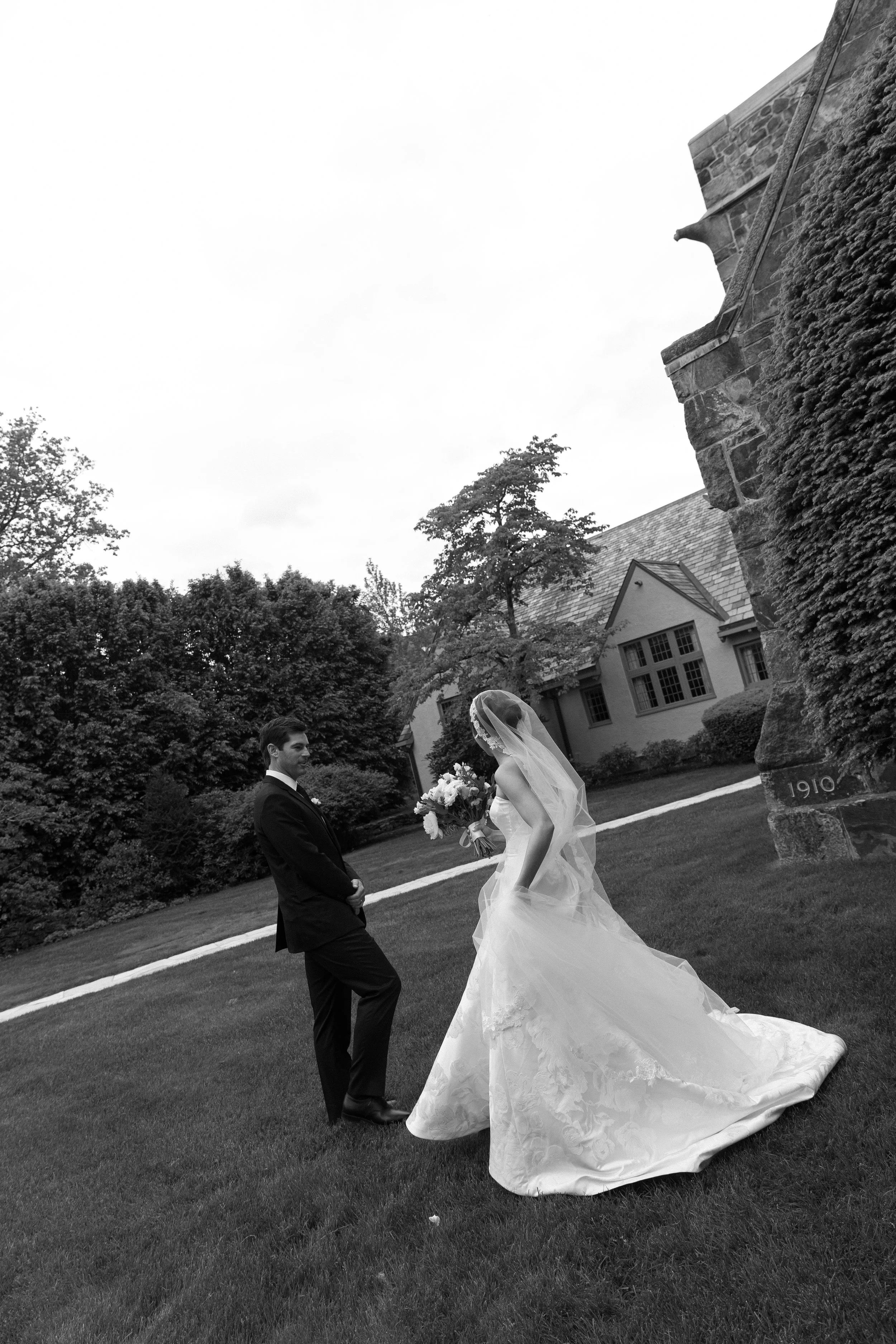 Black and white photo of a bride and groom outdoors on a grassy lawn with trees and a church building in the background. The bride is holding a bouquet and wearing a wedding dress with a veil, while the groom is in a suit.
