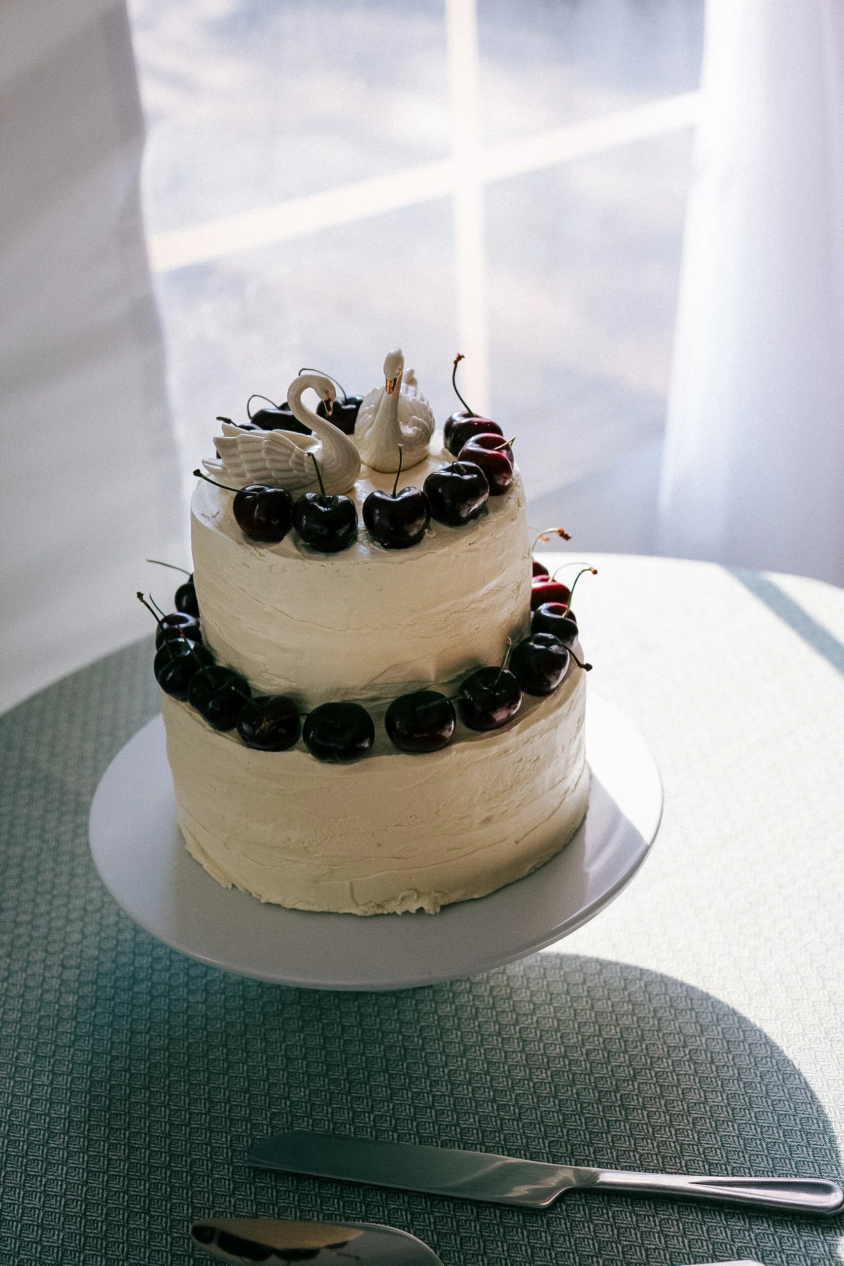 A two-tiered white frosted cake decorated with cherries and swan-shaped toppers, placed on a white cake stand on a table with a knife and spoon nearby.