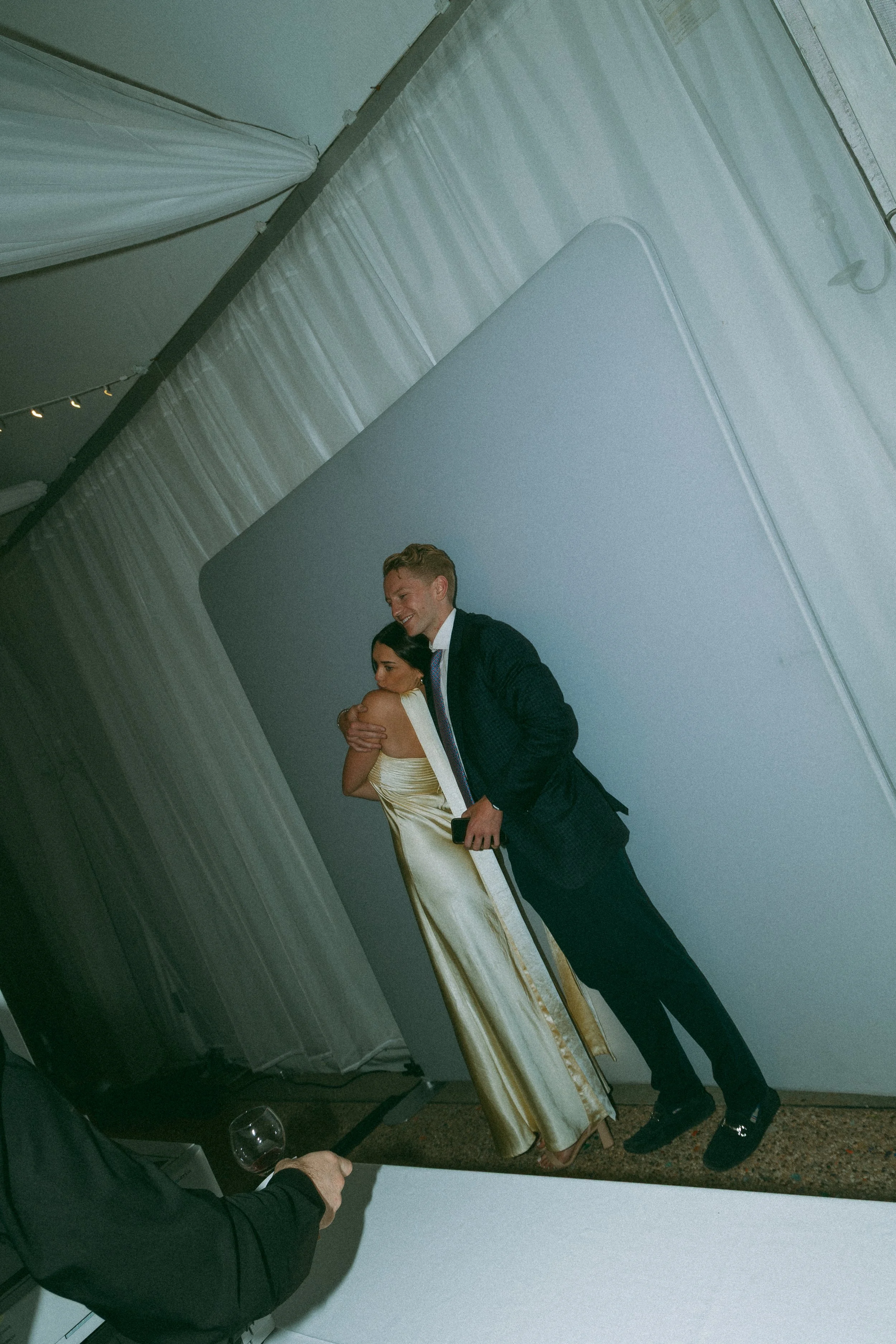 A man in a suit and a woman in a gold dress hugging at an event with white draped curtains and a large white backdrop.