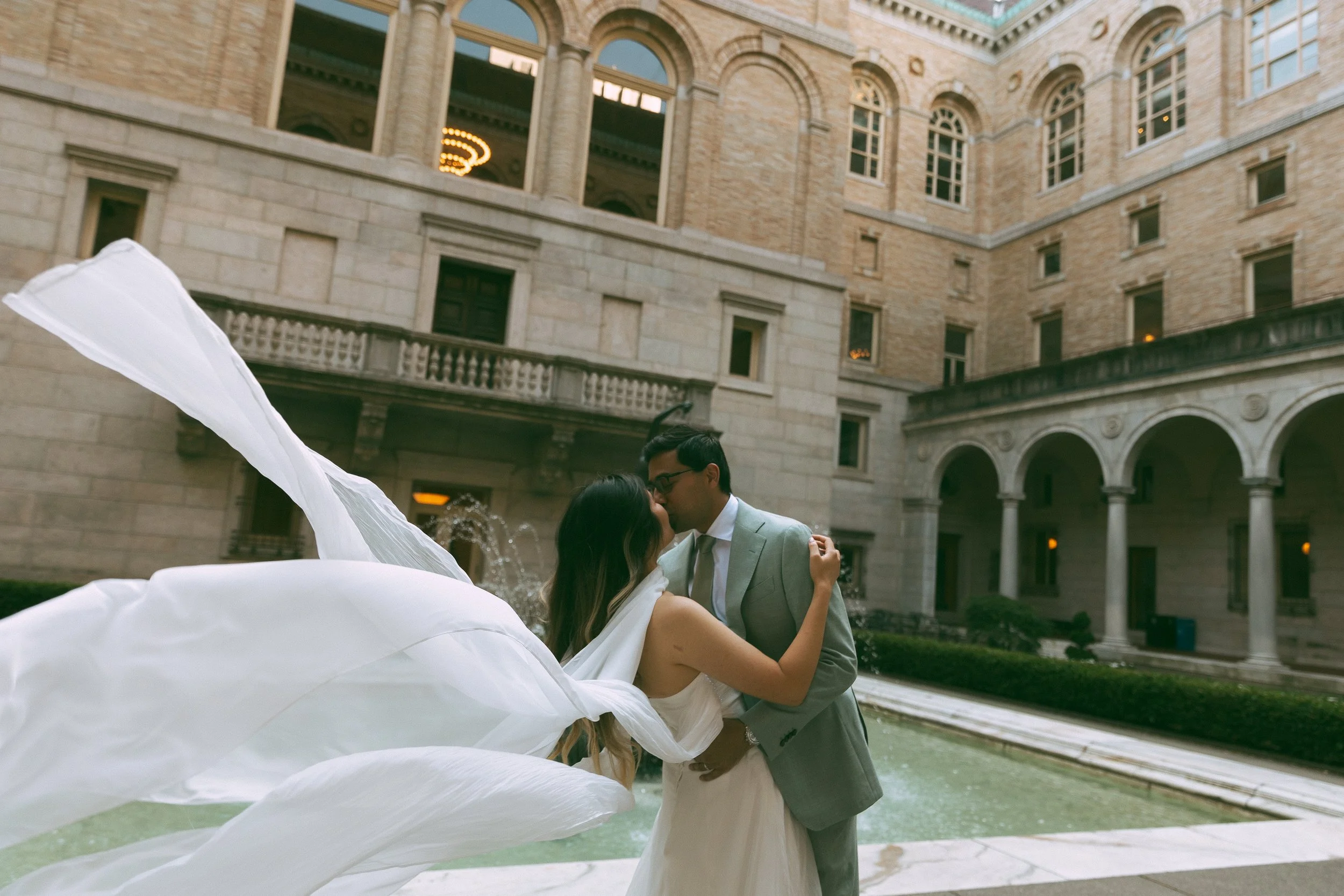 A bride and groom sharing a kiss in front of a fountain in an elegant, historic building courtyard.