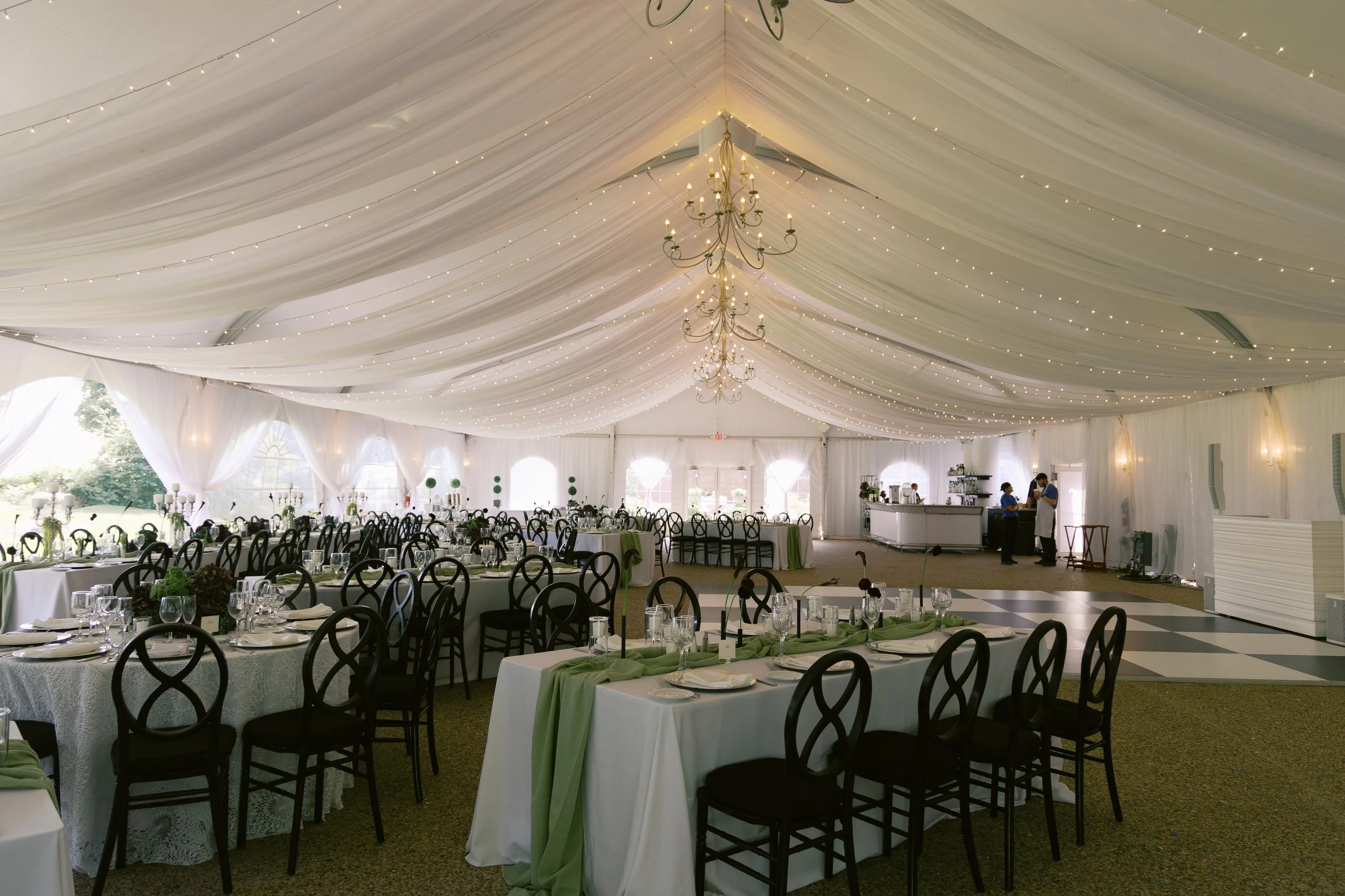 Elegant event tent decorated with string lights, chandeliers, and white drapery, set up with round and rectangular tables with white tablecloths, black chairs, and green table runners for a formal gathering or wedding reception.