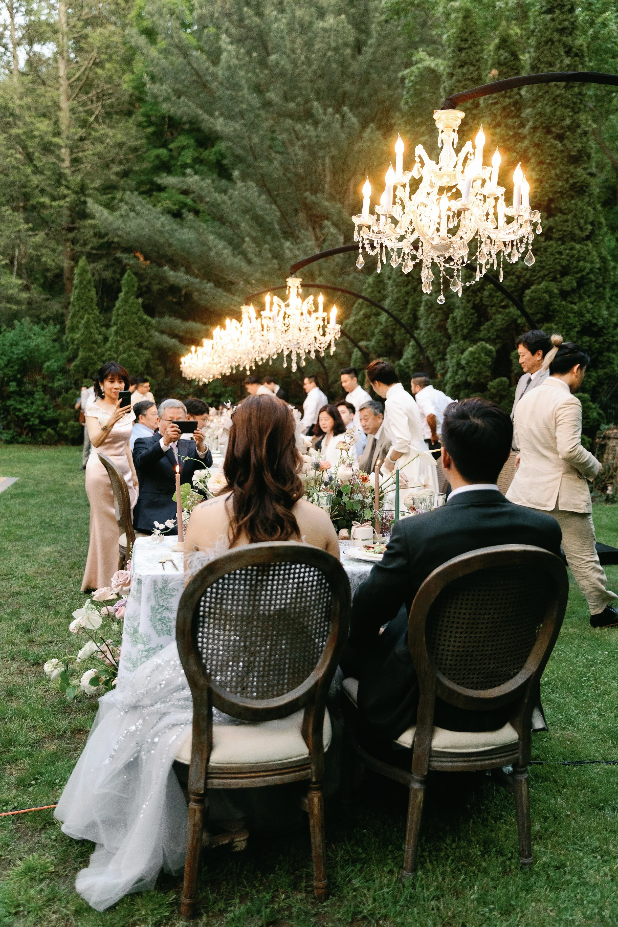 A wedding reception in an outdoor garden setting with underlit chandeliers and guests, including a bride and groom, seated at a table with floral arrangements, surrounded by friends and family, some taking photos.