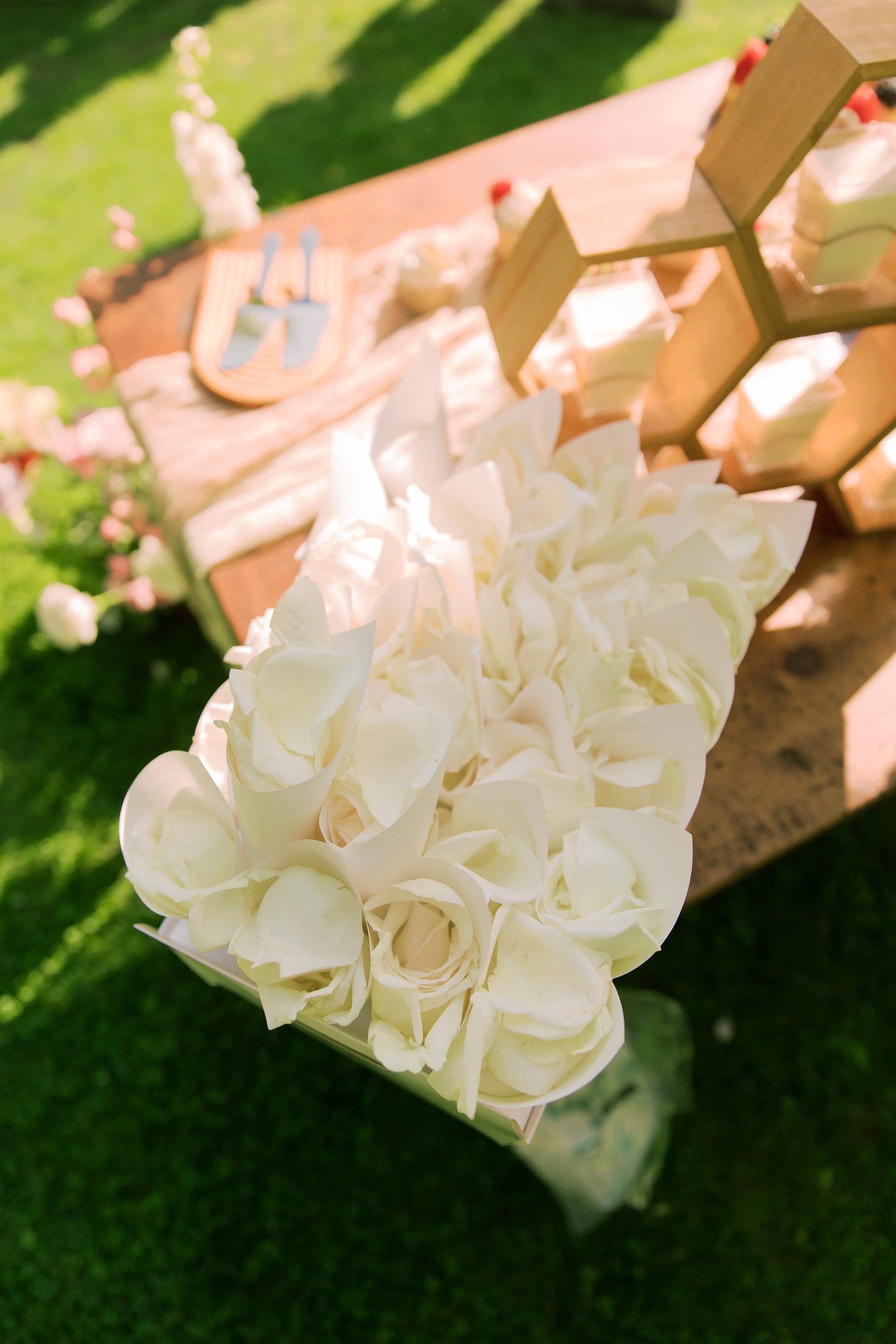 Wedding decor with a flower arrangement of white roses on a table, set outdoors with greenery and pink flowers nearby.