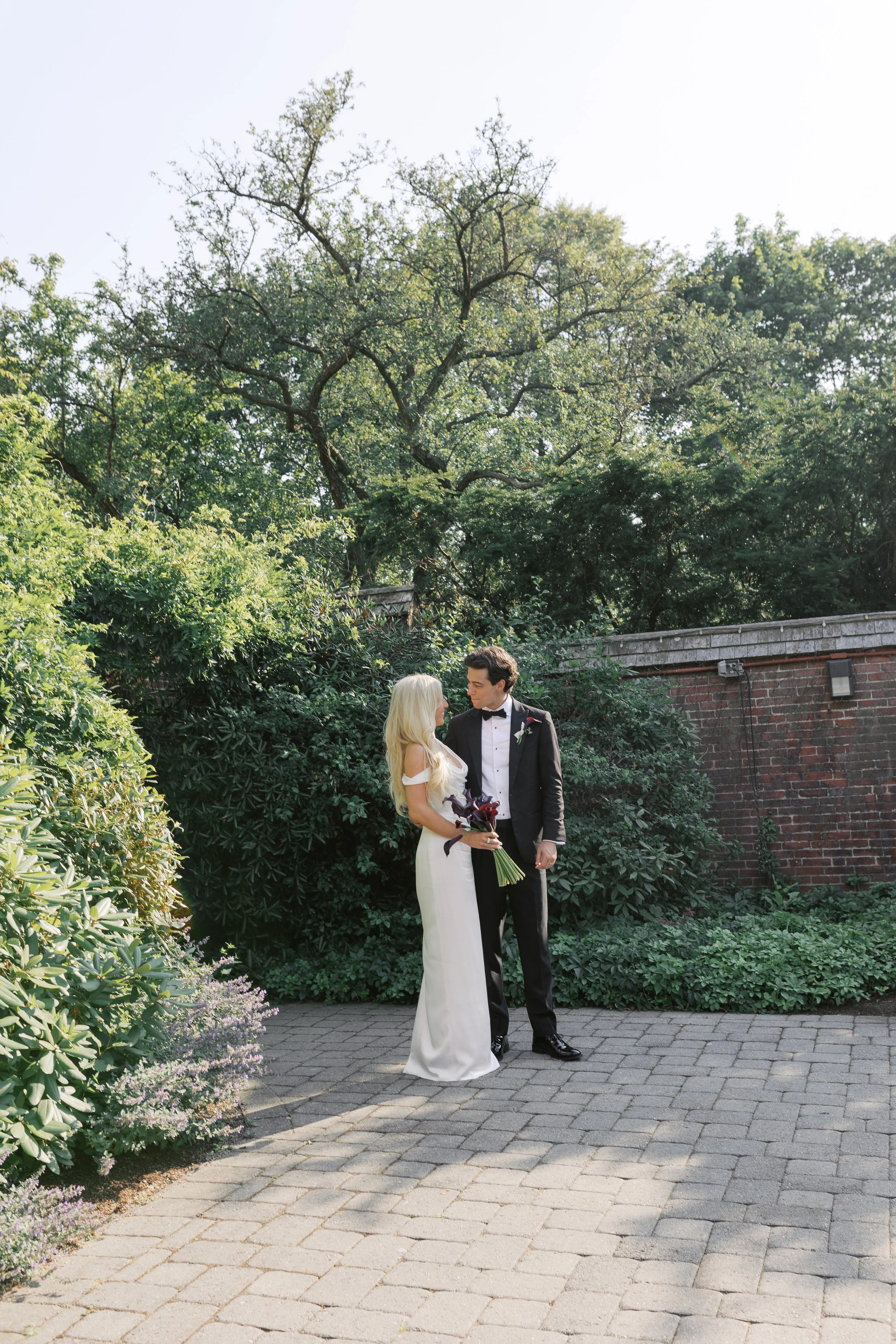 A bride and groom standing outdoors on a paved pathway, with the bride holding a bouquet, surrounded by greenery and a brick wall, during daytime.