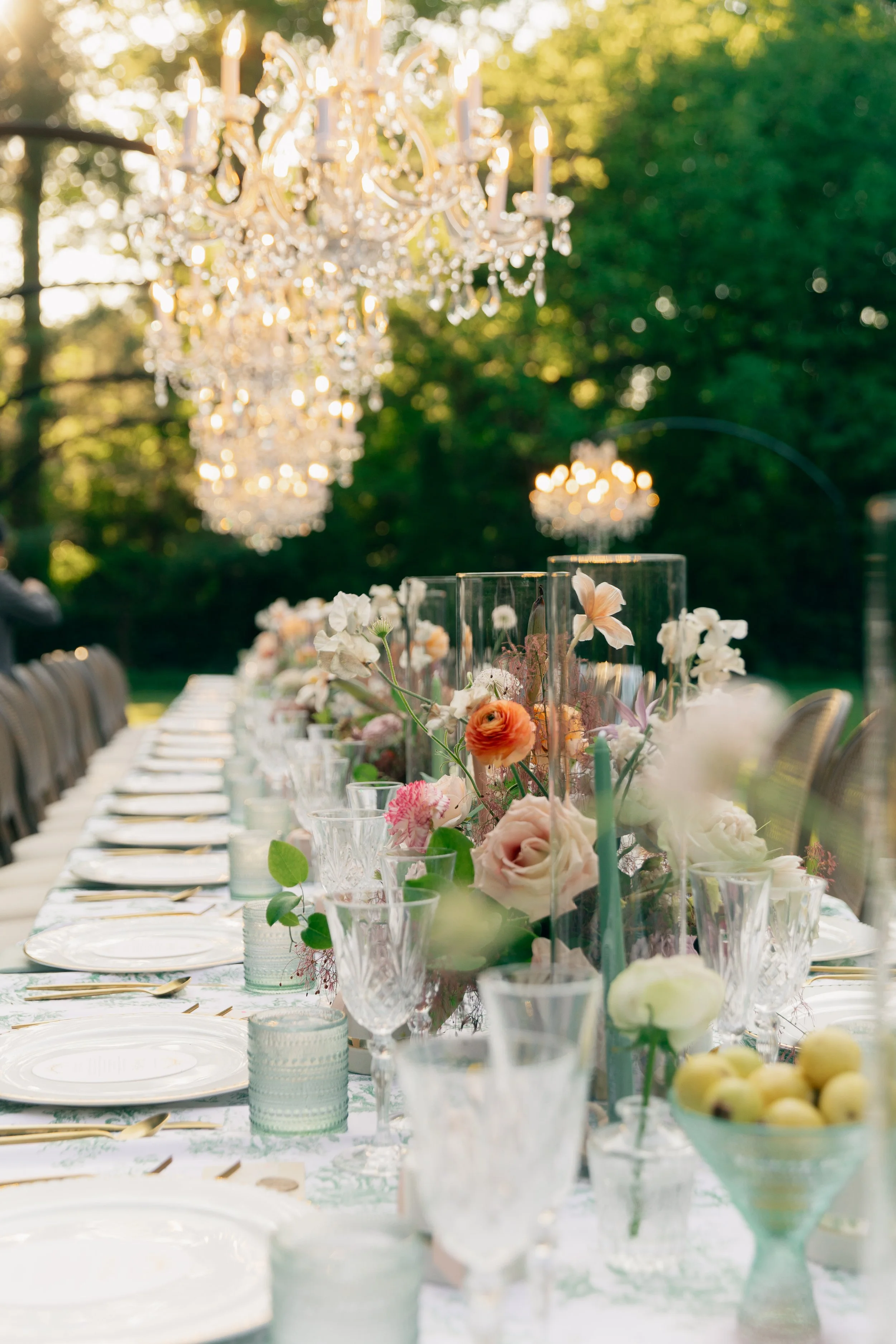 Elegant outdoor dining table with floral centerpieces, fine china, crystal glassware, and chandeliers hanging above in a lush green setting.