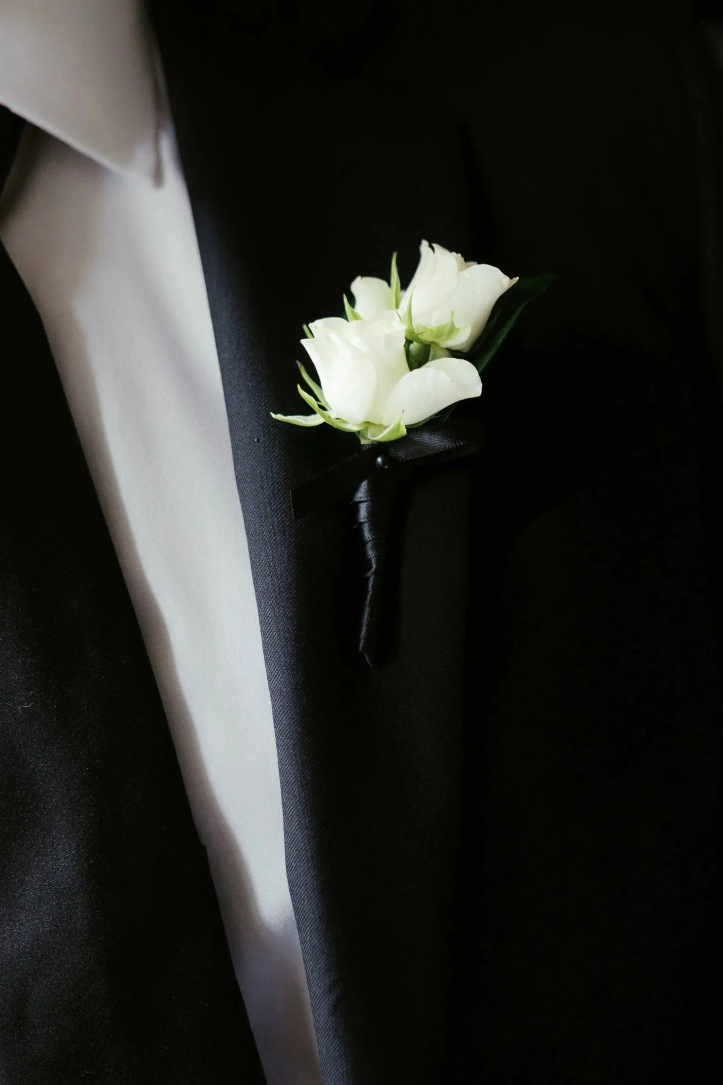 Close-up of a white boutonniere pinned to a black tuxedo lapel, with a white dress shirt visible underneath.
