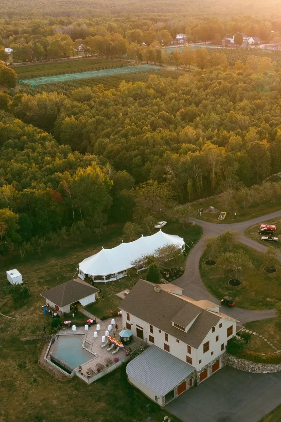 Aerial view of a residential property with a large white event tent, a swimming pool, and multiple vehicles parked on a curved driveway surrounded by trees and greenery at sunset.