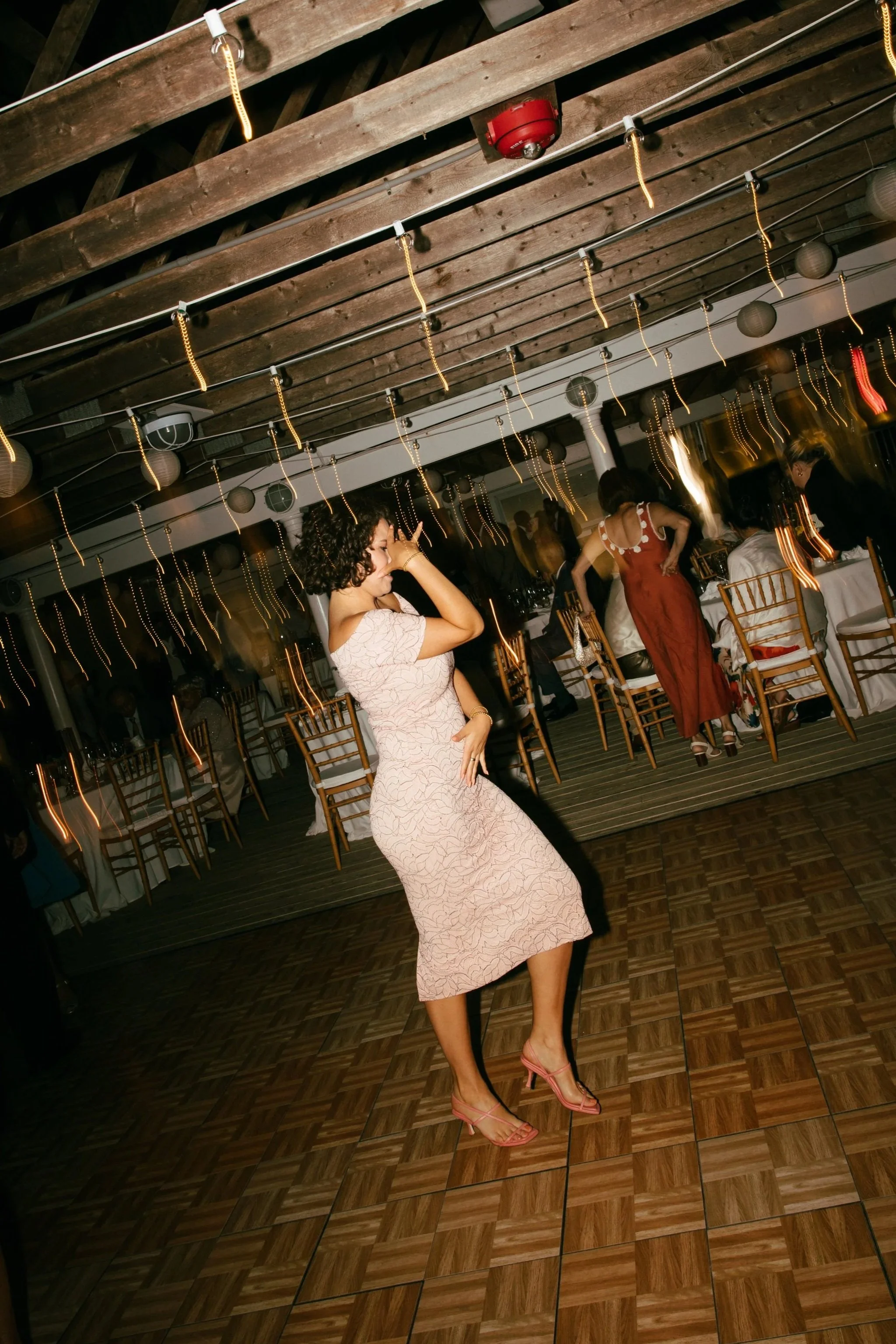 A woman dancing at a party or celebration, wearing a light-colored dress and pink heels, with a lively atmosphere with string lights and guests seated at tables in the background.