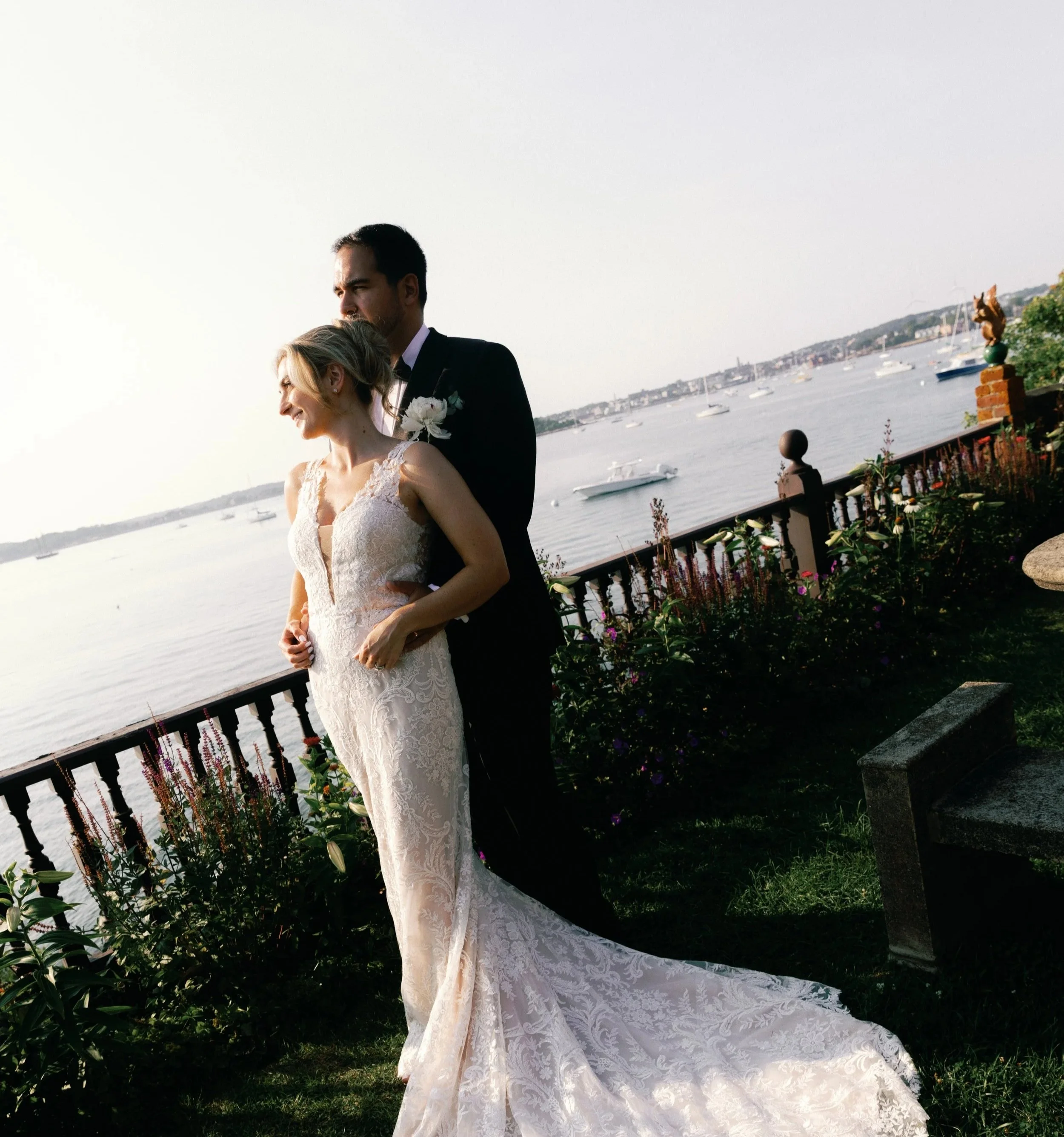A bride and groom standing together on a grassy area near a water body with boats, against a backdrop of a clear sky and a distant shoreline.
