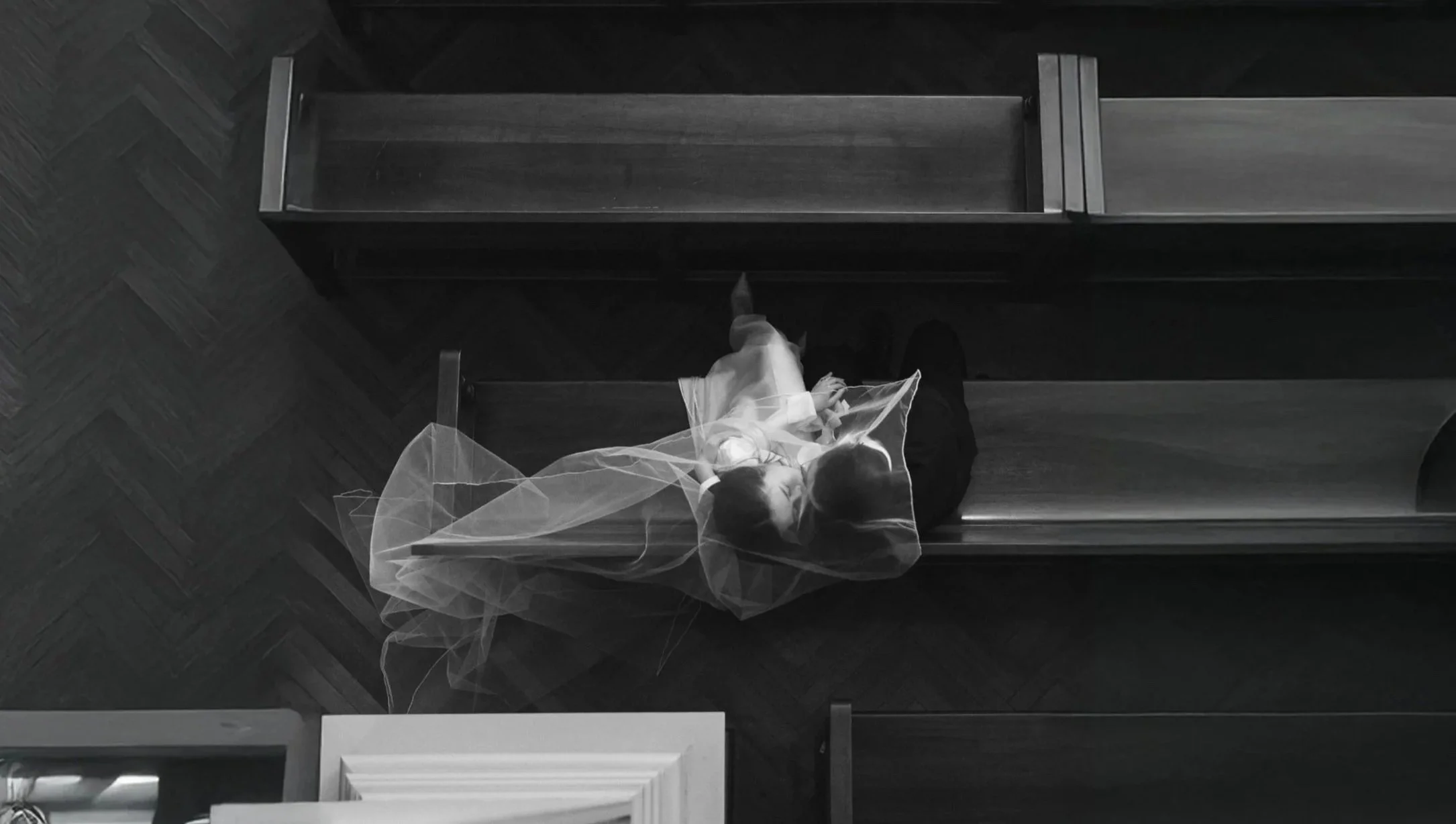 A girl wearing a veil is sitting alone on a wooden church bench, looking up, with an overhead view in black and white.