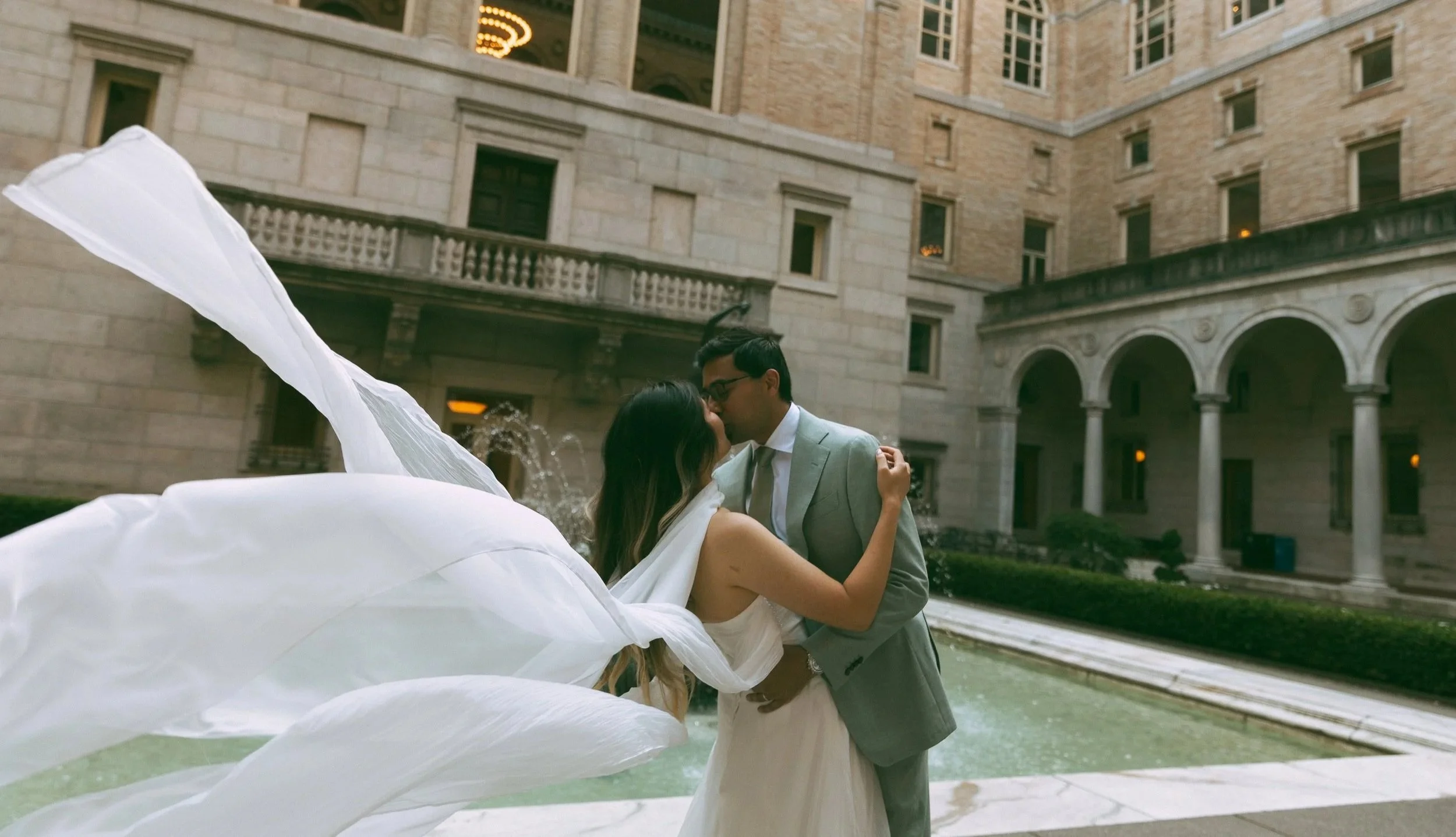 A bride and groom sharing a kiss in front of a fountain in an elegant, historic building courtyard.