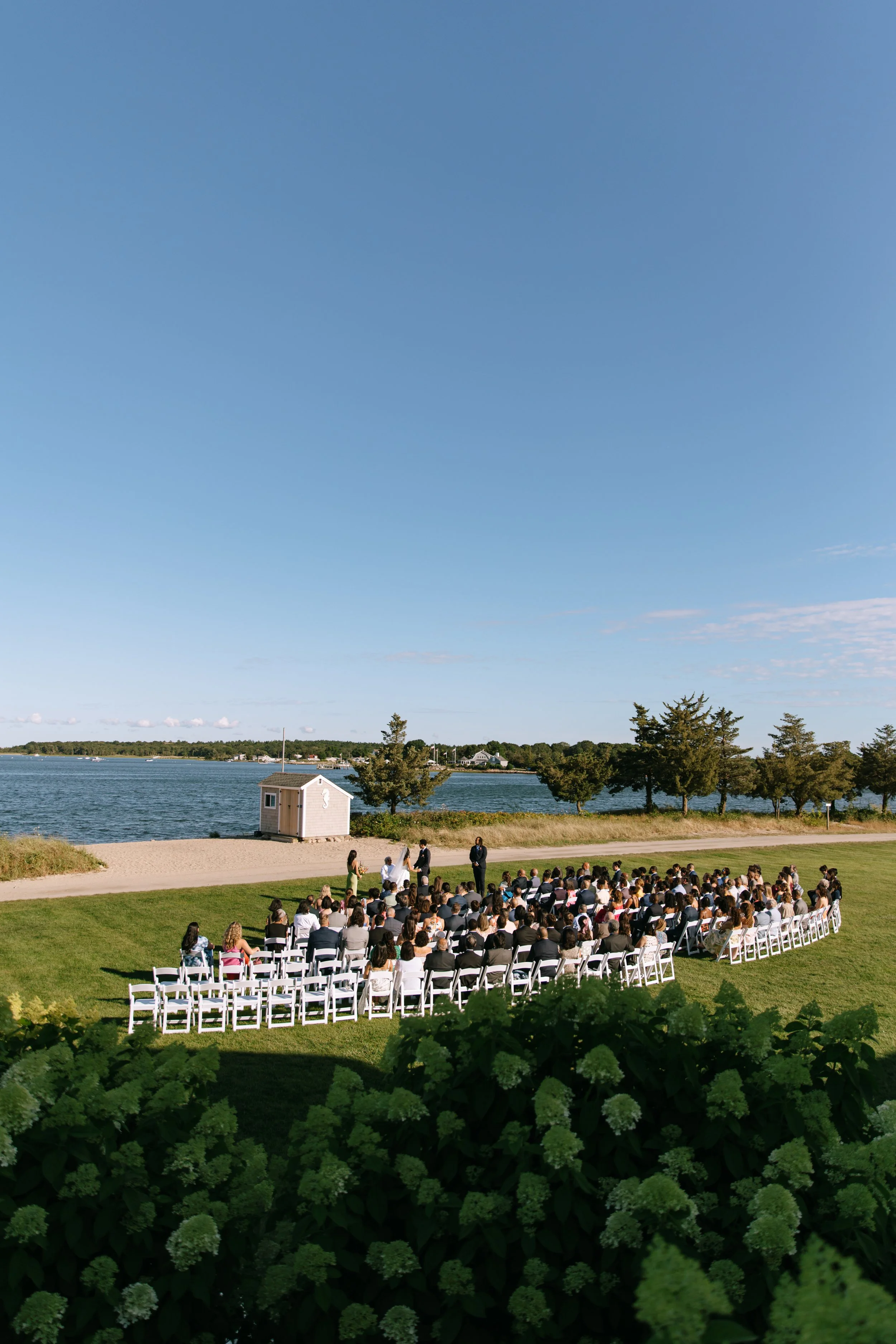 Outdoor wedding ceremony taking place on a grassy area near a body of water, with guests seated on white chairs and a small group of people standing near the officiant.