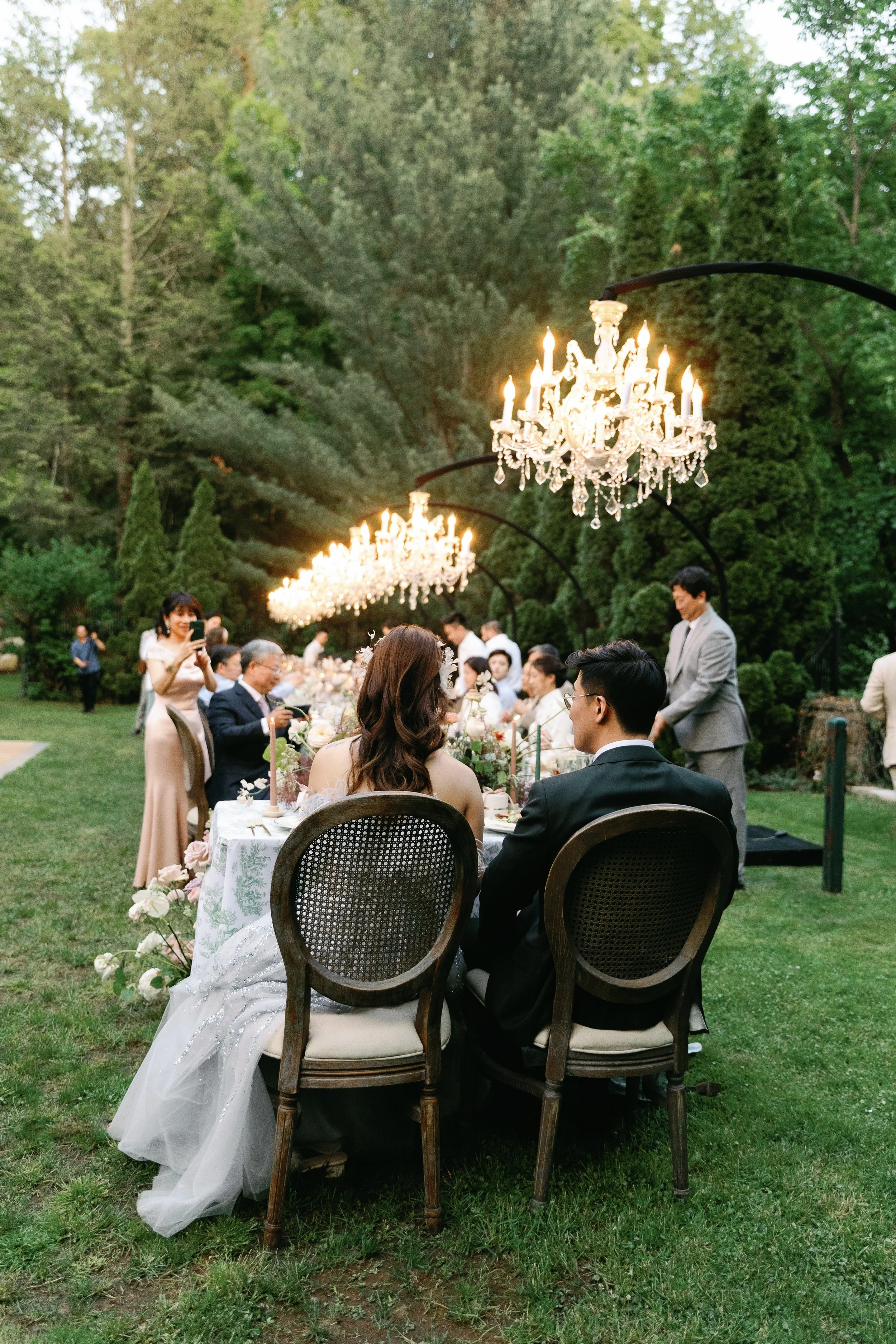 A wedding reception outdoors with a long dining table decorated with flowers, chandeliers hanging overhead, and guests seated around enjoying the meal, surrounded by trees and greenery.