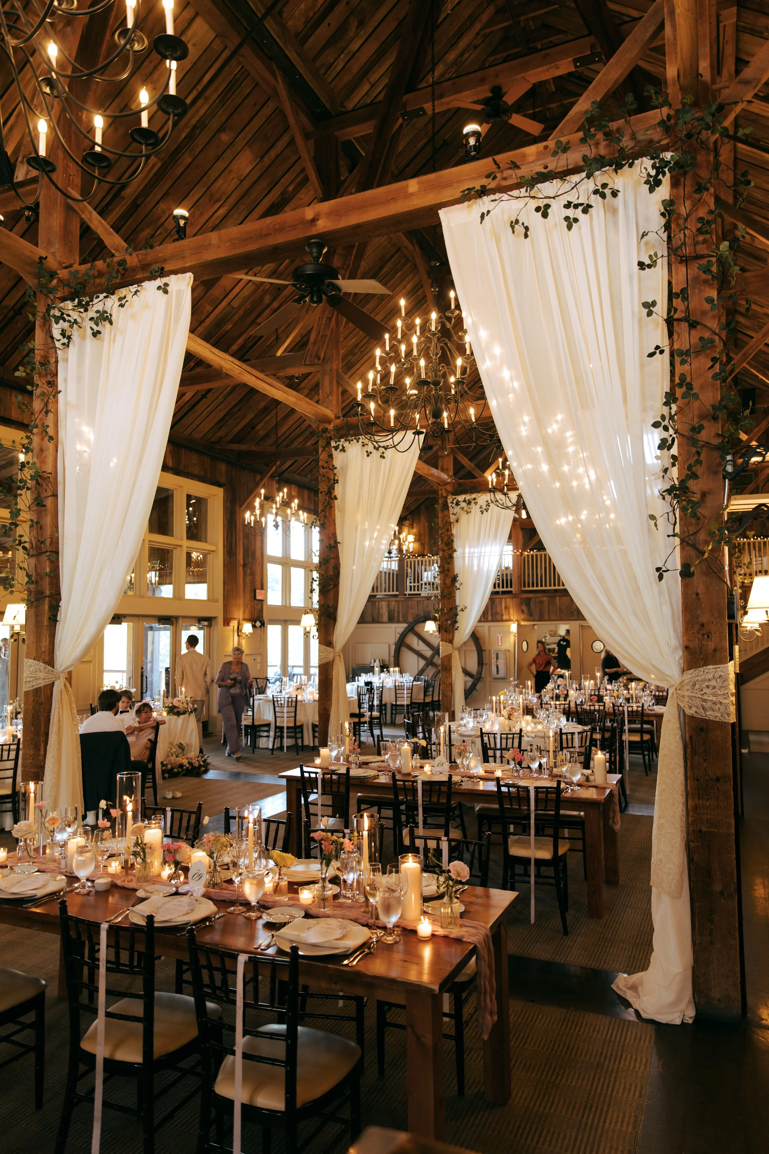 Elegant rustic wedding reception with wooden beams, white draped curtains, chandeliers, and tables set with candles, flowers, and fine tableware.