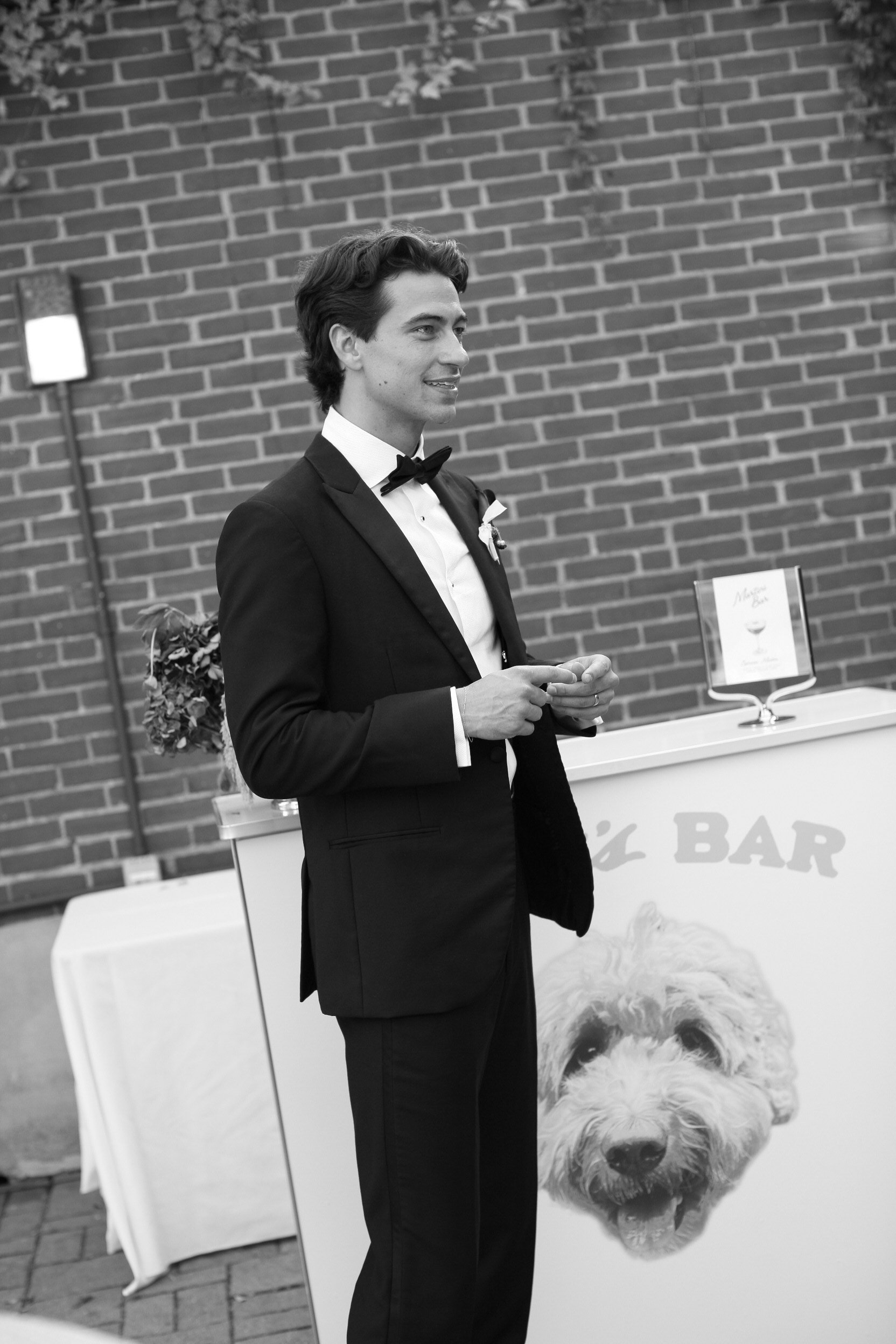 A man dressed in a tuxedo with a bow tie standing in front of a brick wall, holding a small object in his hands. There is a sign with a dog's face and the words 'Dog Bar' on it in the background.