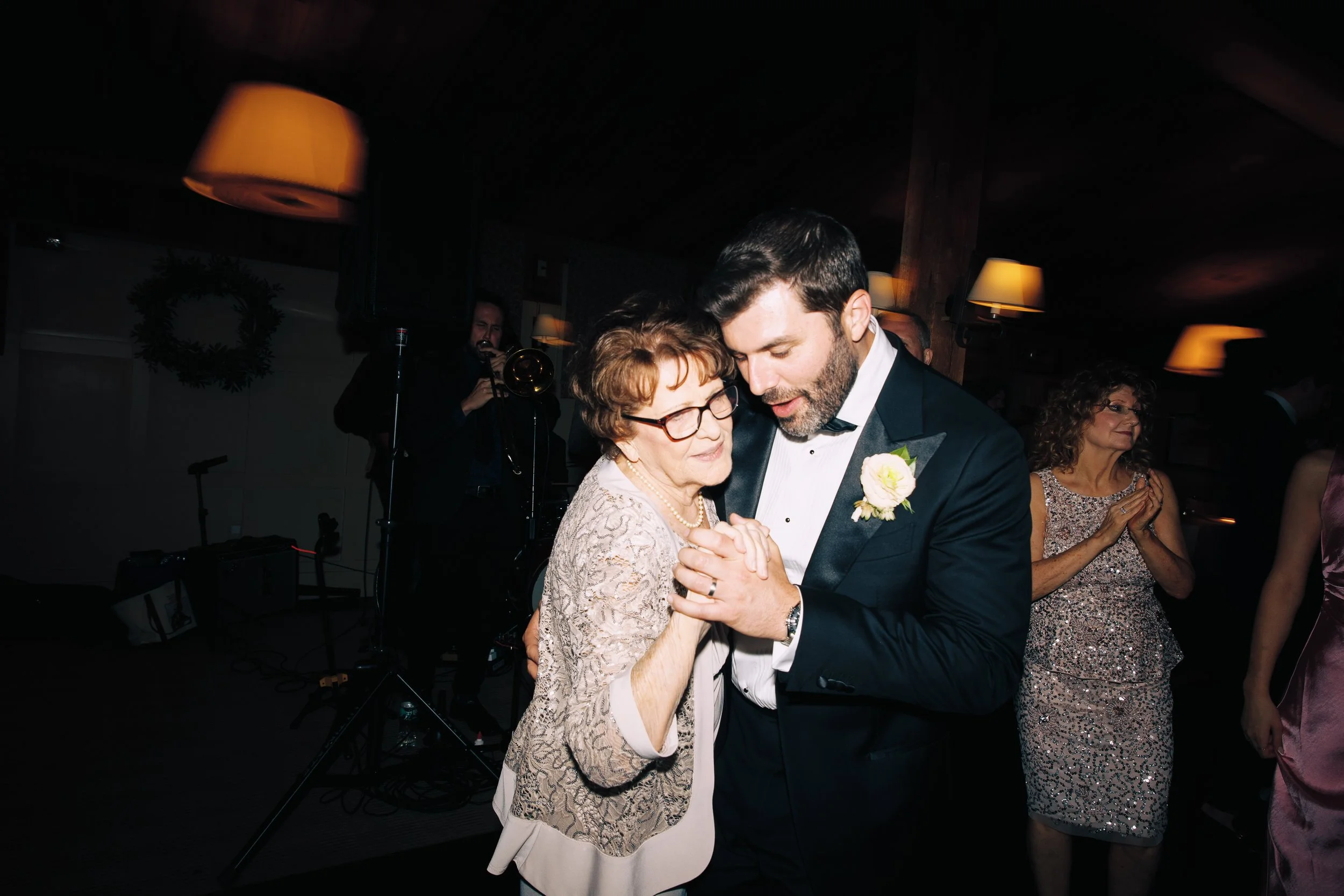 A man in a tuxedo dancing with an elderly woman at a celebration or wedding reception, holding hands and close to each other, with other guests and a band in the background.