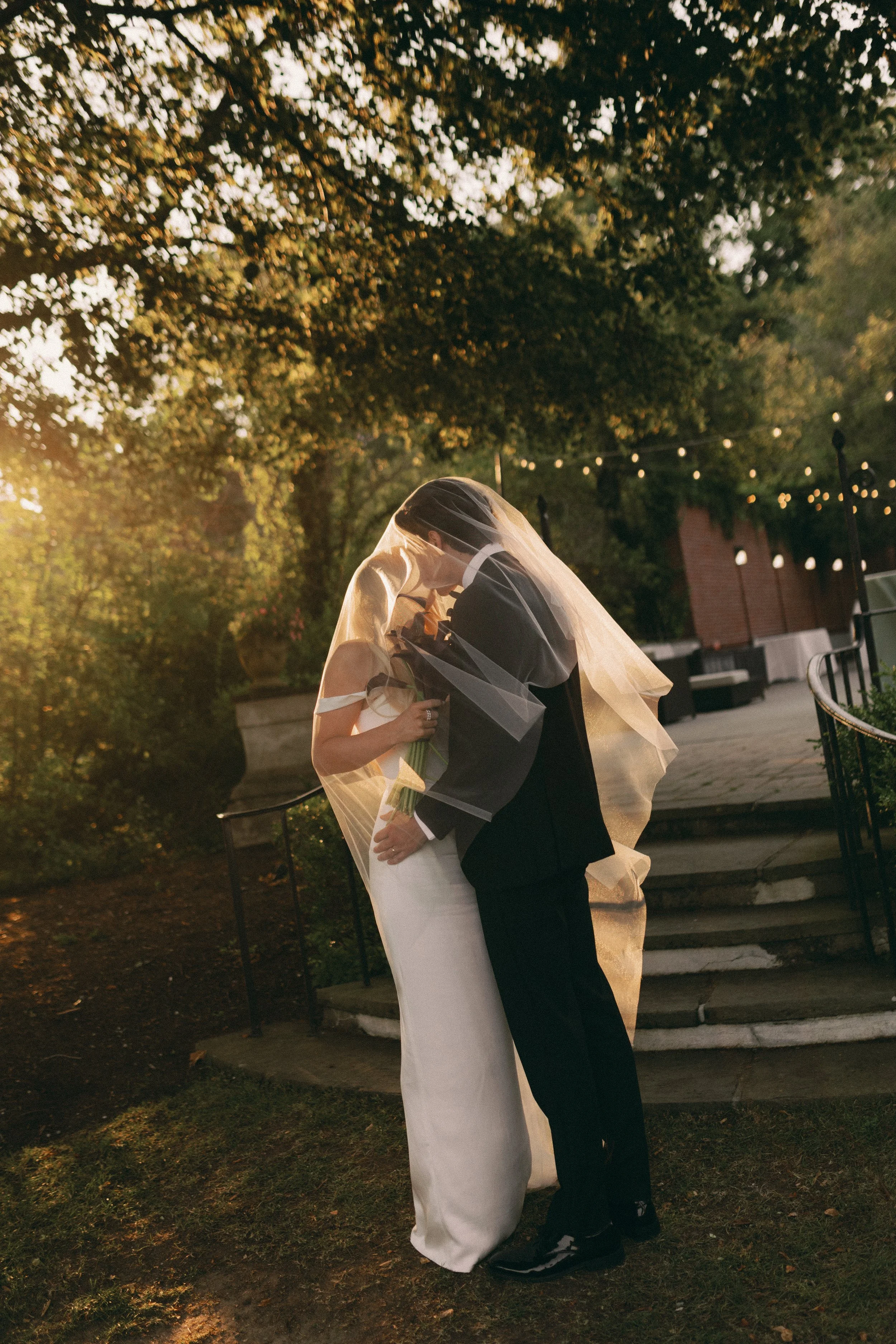 A bride and groom sharing a kiss outdoors during sunset, with the bride holding a bouquet and both dressed in wedding attire, framed by trees and string lights.