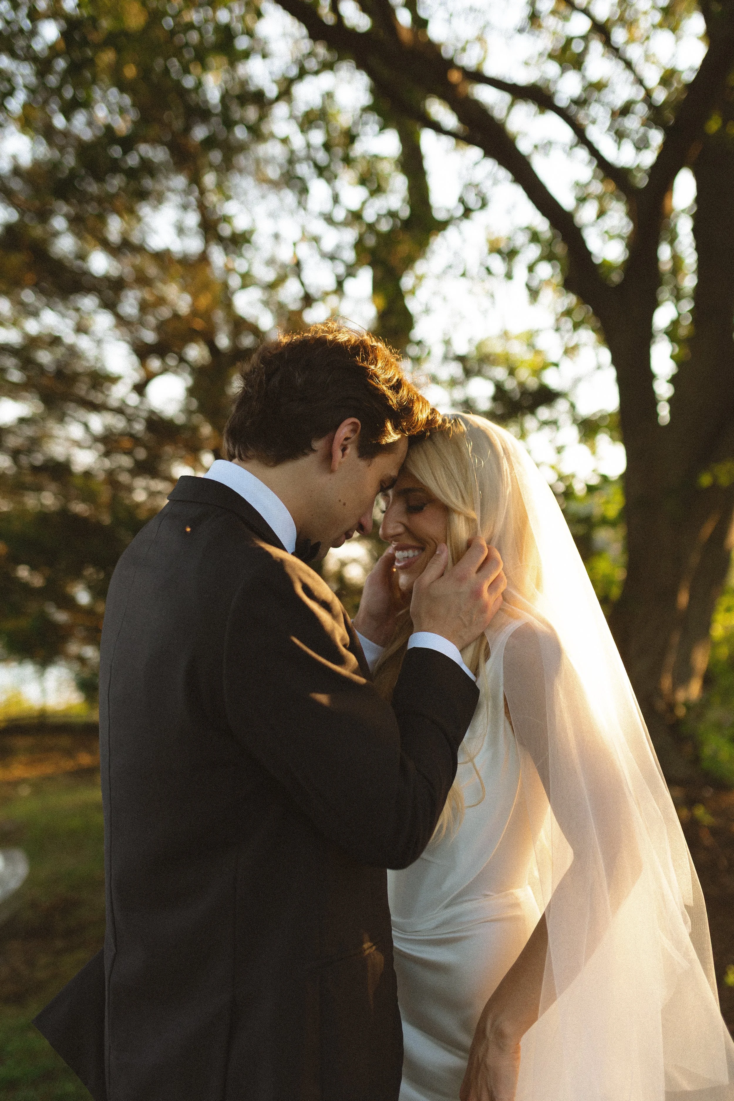 A bride and groom share a tender moment outdoors during sunset, with trees and fall leaves in the background.