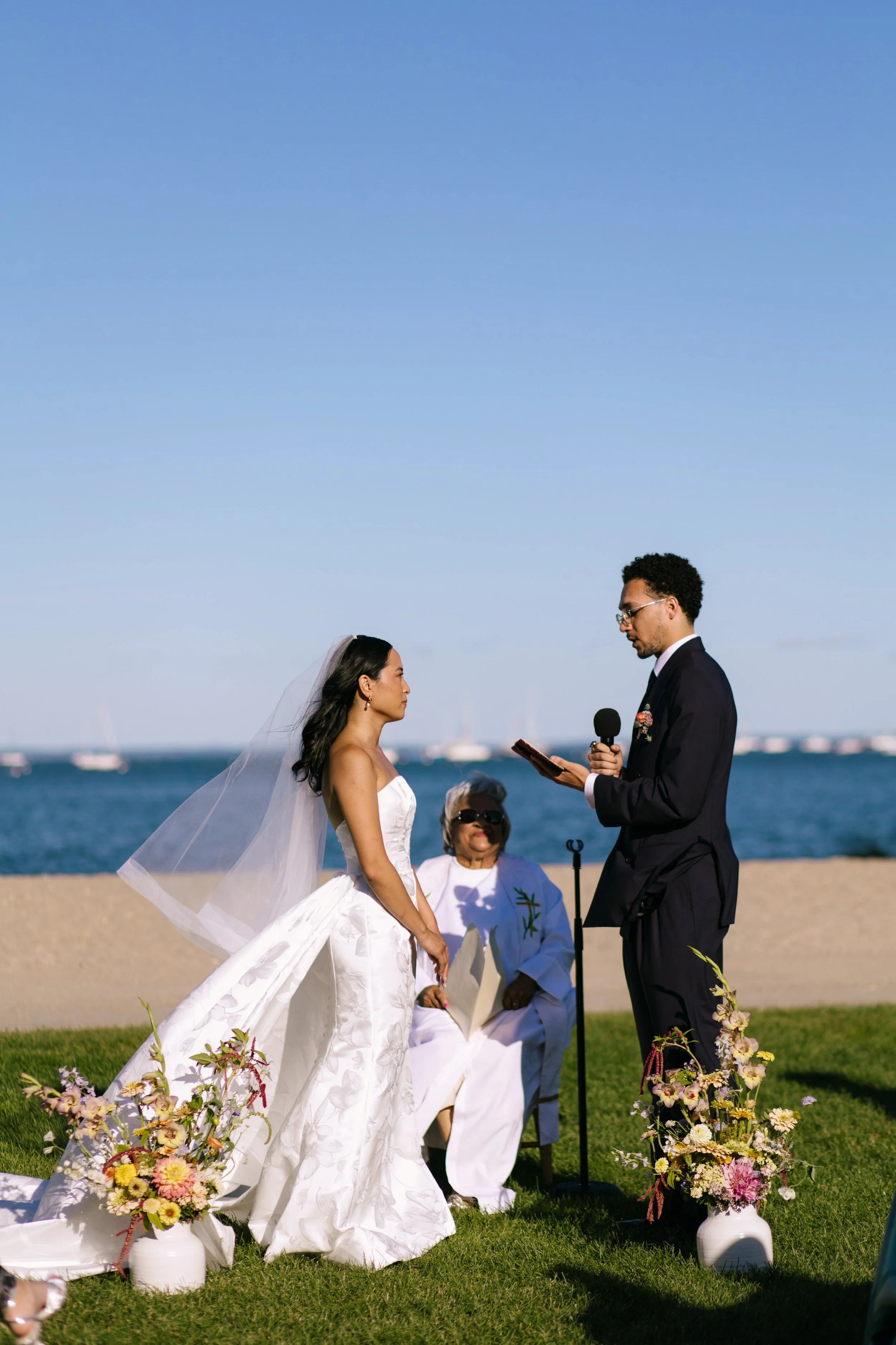 A couple is exchanging wedding vows outdoors near the water, with a woman officiant seated behind them, under a clear blue sky. The bride is in a white strapless gown with a veil, and the groom is in a dark suit holding a microphone and reading vows.
