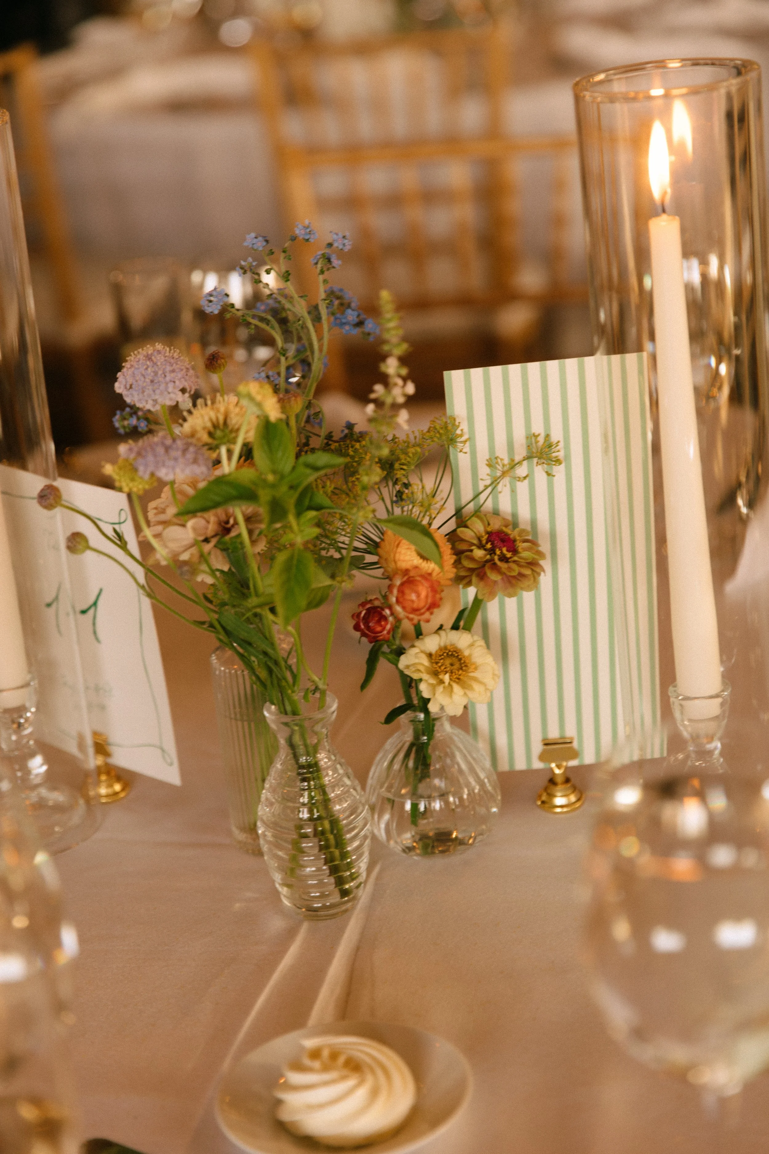 A decorative table centerpiece featuring glass vases with mixed fresh flowers, striped place cards, and tall candles in glass holders, set on a beige tablecloth.