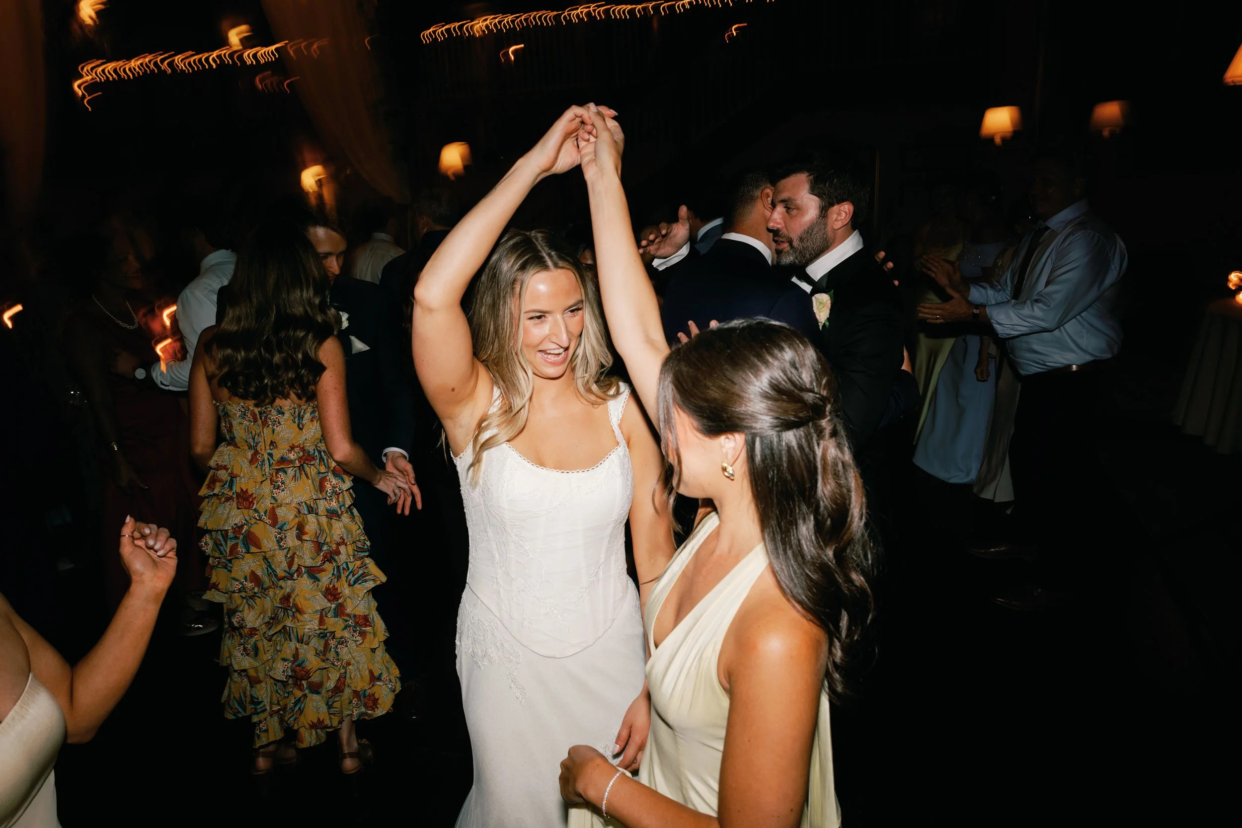 A woman in a white wedding dress dancing with a woman in a cream dress at a wedding reception, with other guests dancing in the background.