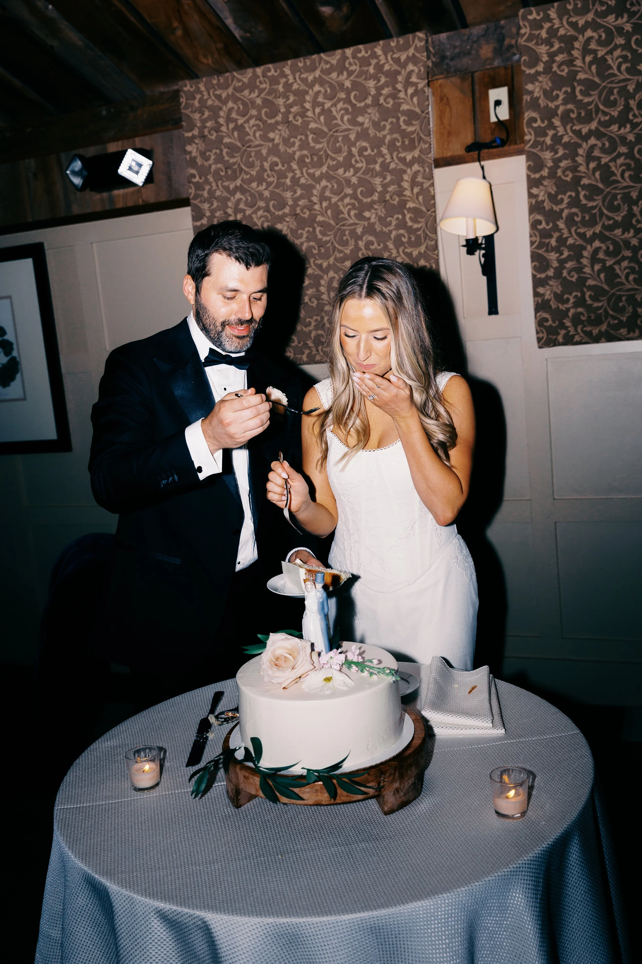 A bride and groom sharing a piece of wedding cake during their celebration. The bride is covering her mouth with her hand, and the groom is holding a fork with cake. A wedding cake decorated with flowers and a figurine is on the table, along with lit