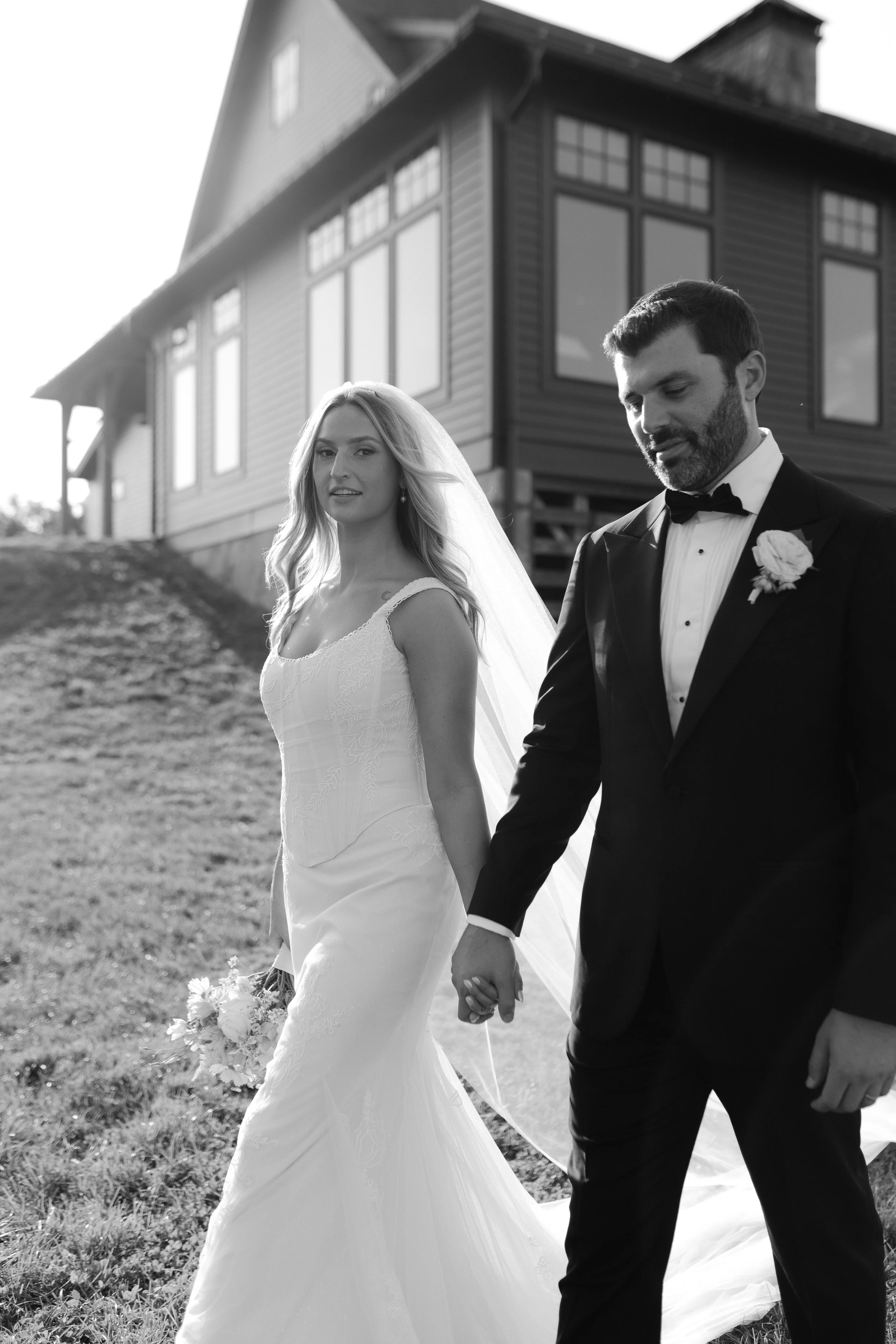 Black and white photo of a bride and groom walking hand in hand outdoors, with a house in the background.