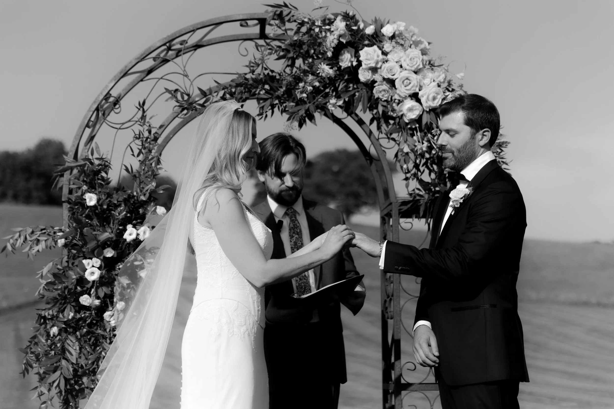 Wedding ceremony outdoors, bride and groom exchanging rings under a floral arch with officiant.