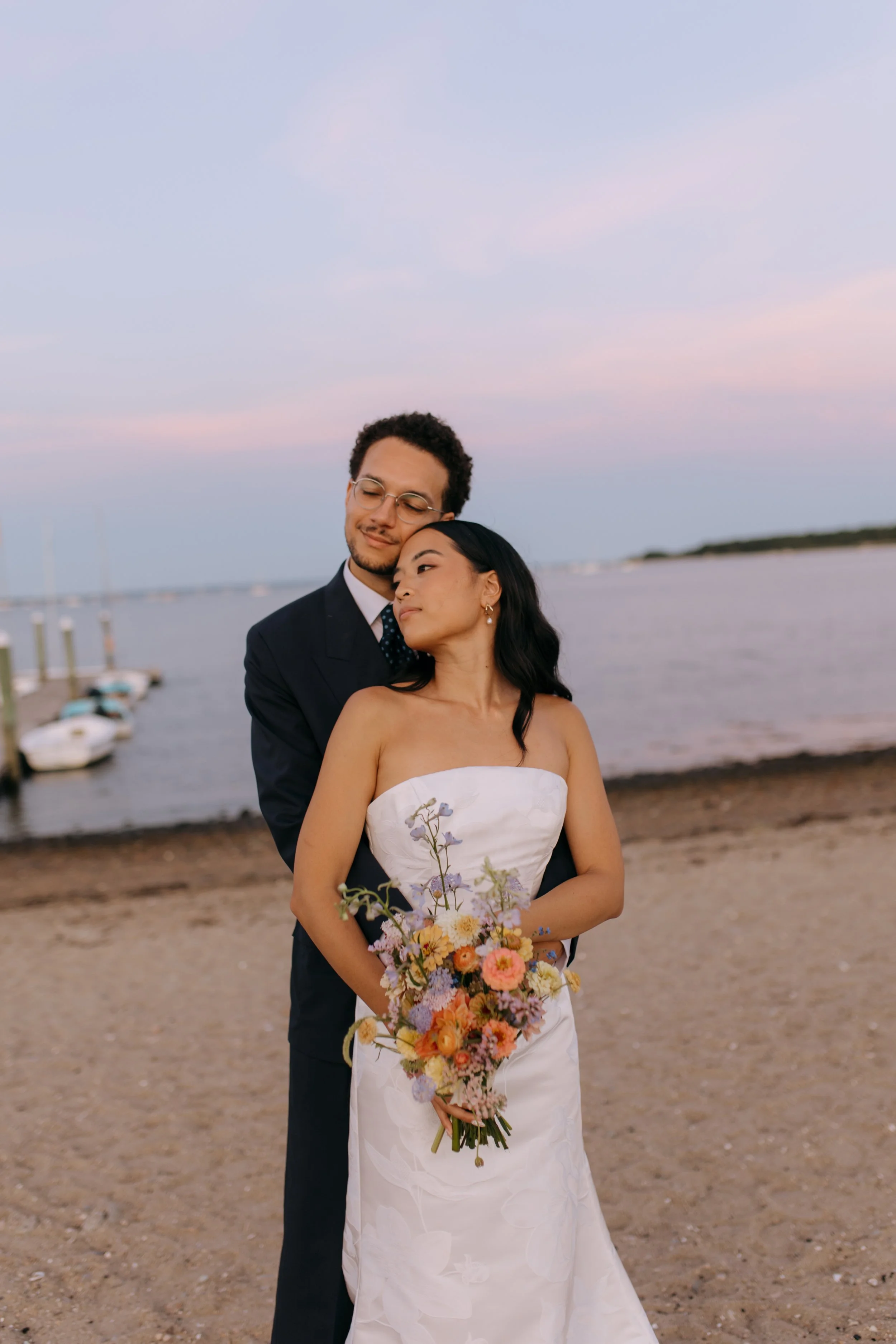 A couple in wedding attire stands on a sandy beach at sunset, with the man embracing the woman from behind, holding a bouquet of colorful flowers.
