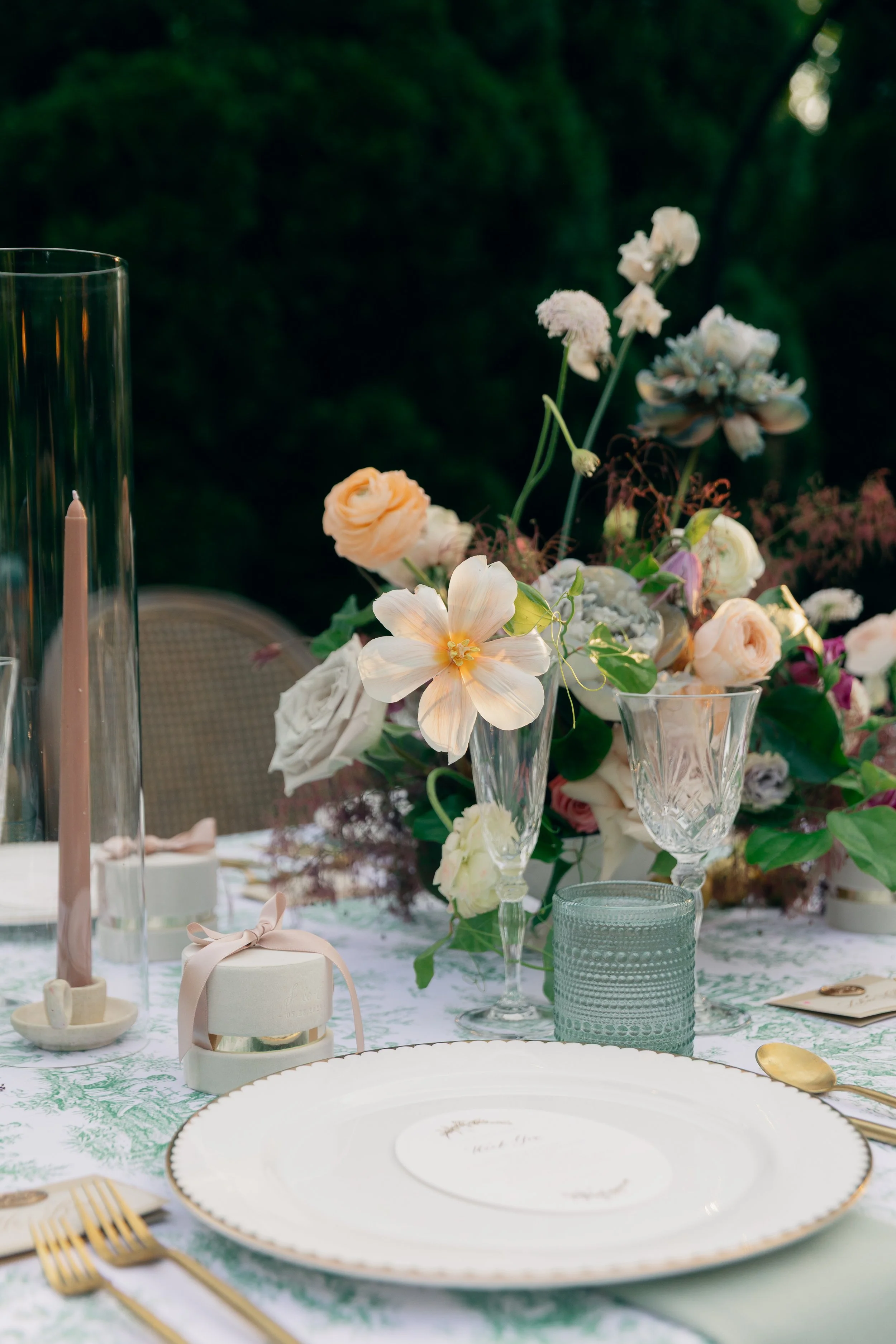 Elegant outdoor table setting with a floral centerpiece, crystal glassware, gold flatware, a pink candle in a glass holder, and a white plate with a small dish on top, all arranged on a white tablecloth with green accents.
