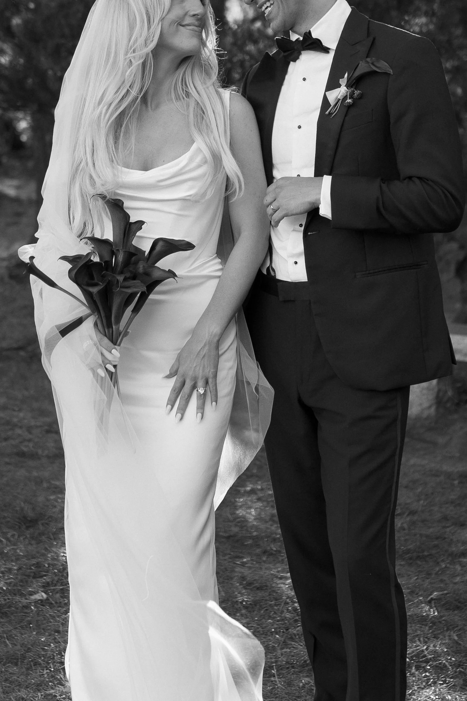 A bride and groom stand close together outdoors, holding hands. The bride wears an elegant white wedding dress with a veil and holds a bouquet of dark-colored flowers. The groom is dressed in a tuxedo with a bow tie, smiling at the bride. In black an