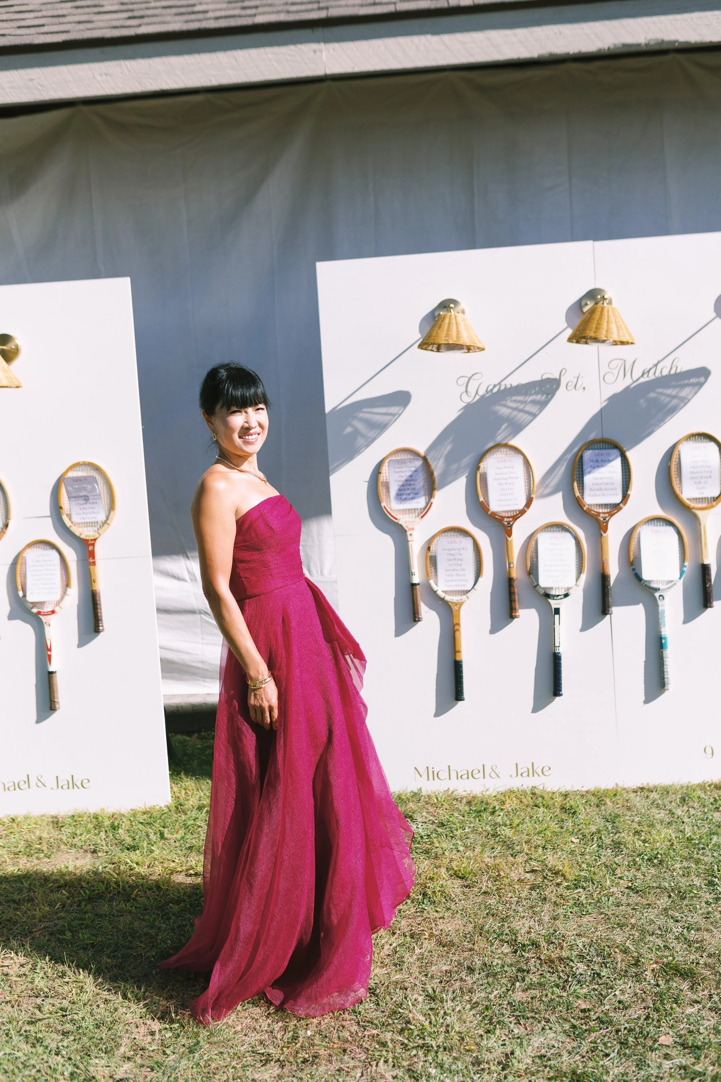 A woman in a strapless magenta dress standing outdoors in front of white display boards decorated with tennis rackets and reading materials, with shadows cast by wall lamps behind her.