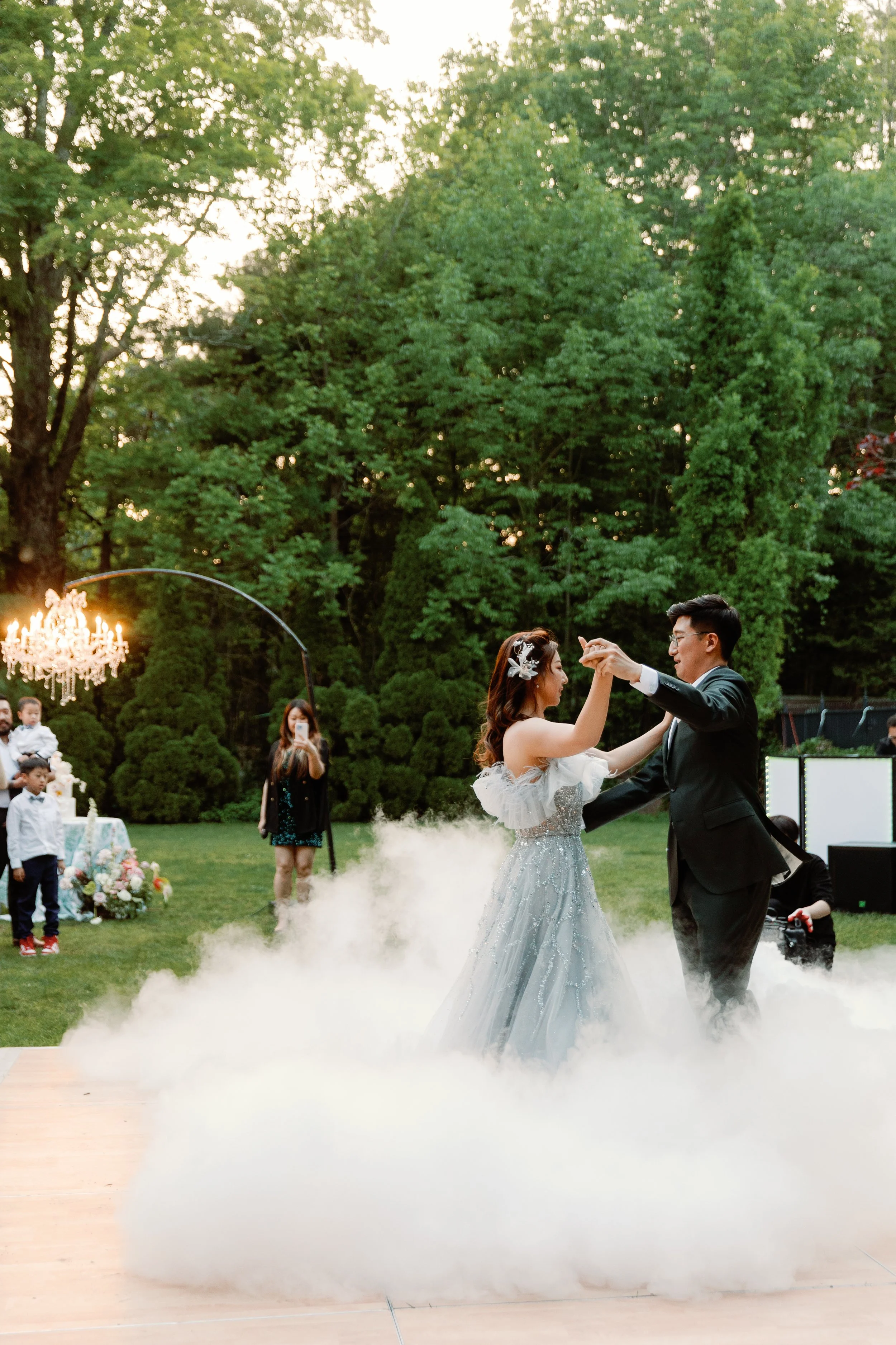 Couple dancing at an outdoor wedding reception, surrounded by green trees and a fog-like effect on the ground.