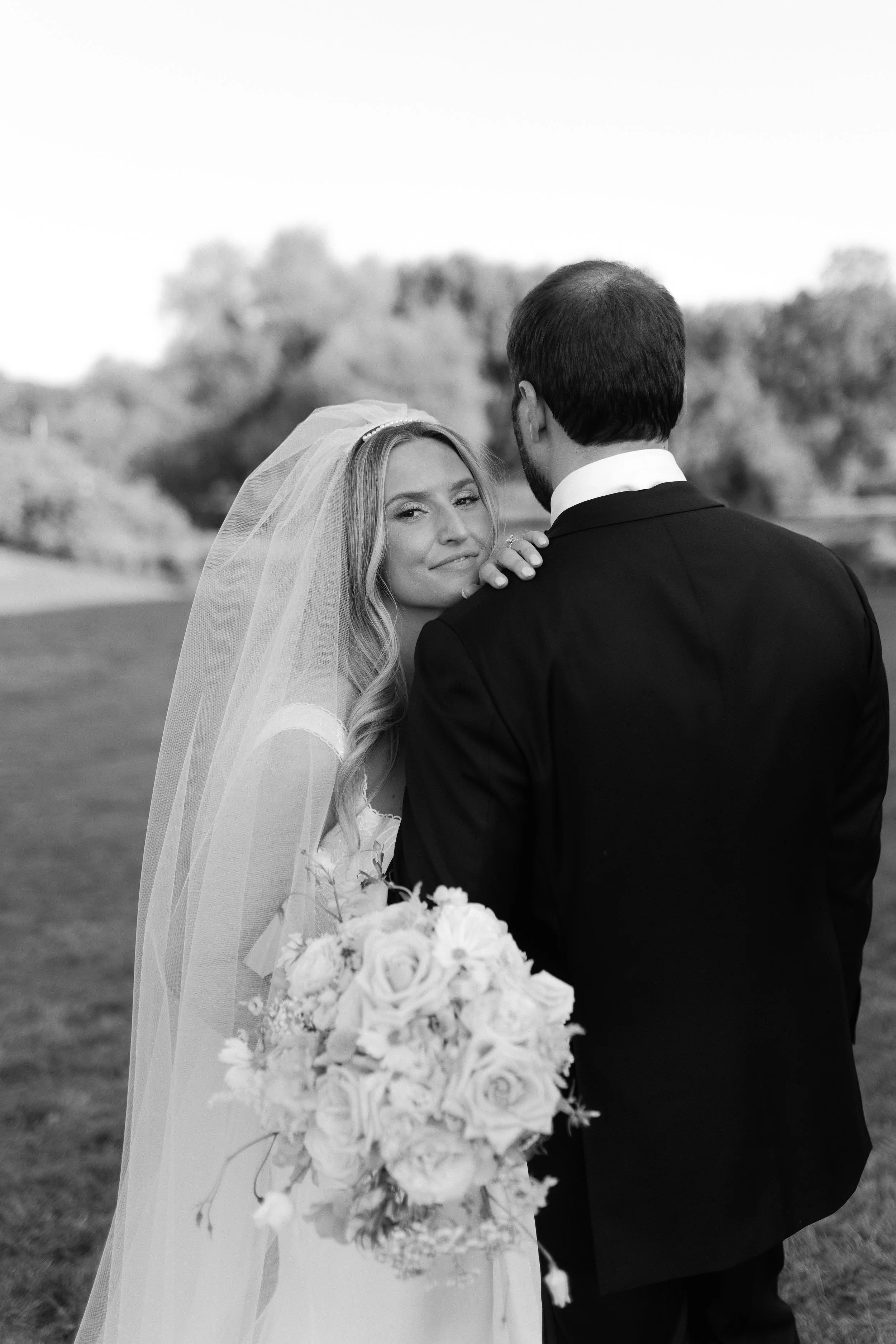 Bride and groom on their wedding day standing outdoors, with the bride holding a bouquet of roses and gazing at the camera.