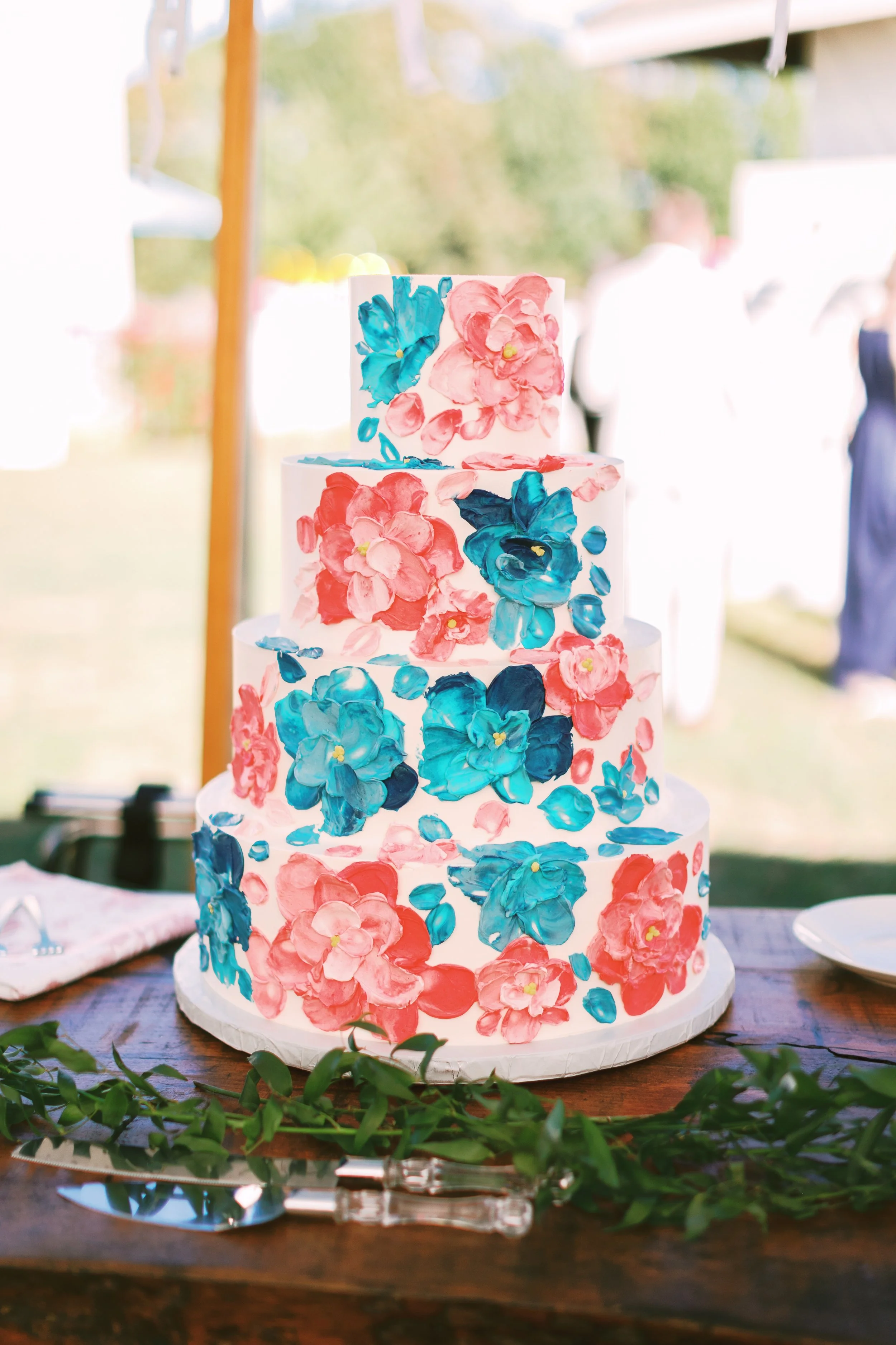 A colorful, multi-tiered wedding cake decorated with pink and blue floral icing, placed on a wooden table with greenery and cake utensils in the foreground.