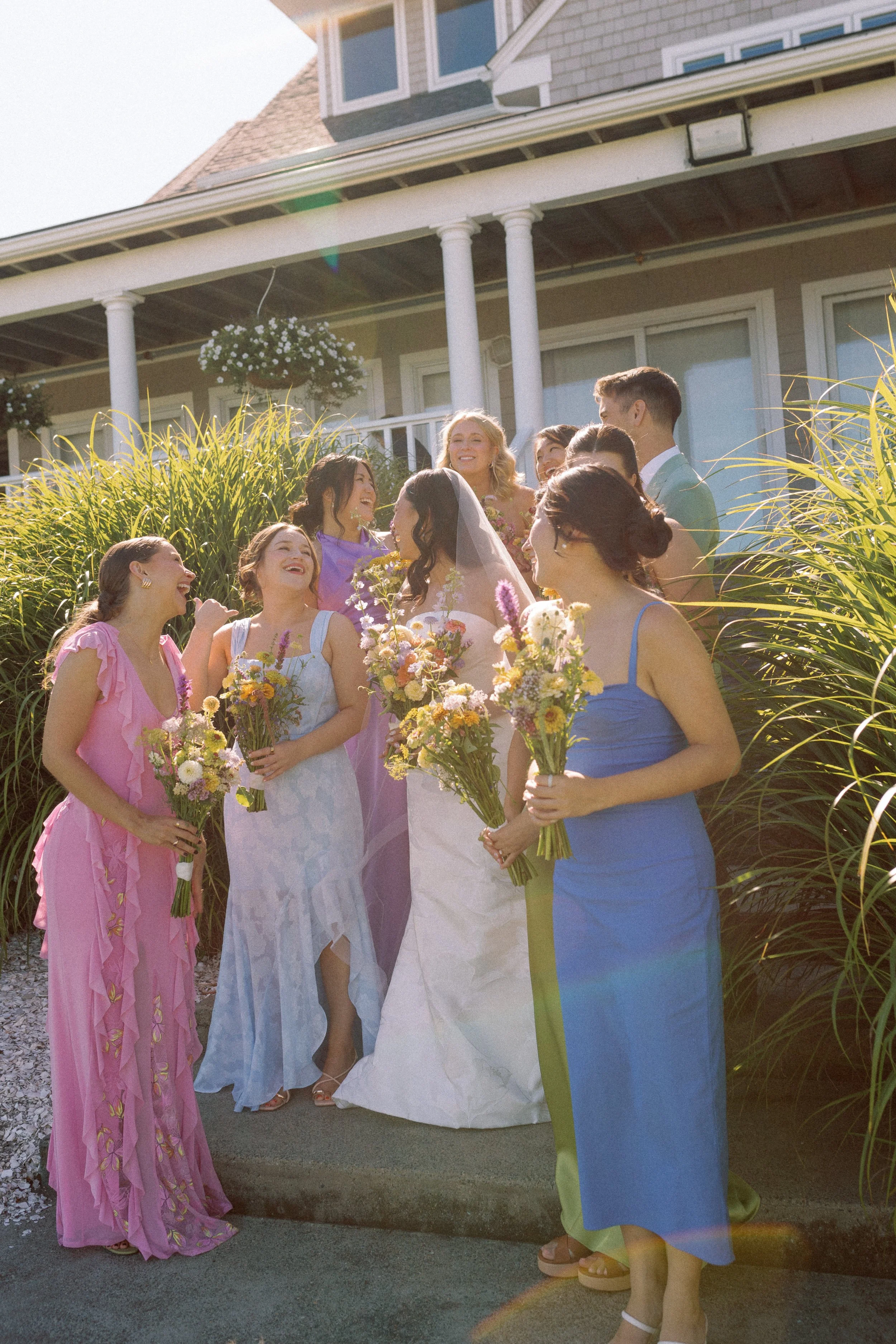 A group of people, including a bride in a white wedding dress and veil, standing outside a house with white pillars and a porch, surrounded by greenery and flowers. They are smiling, holding bouquets, and sharing a joyful moment on a sunny day.